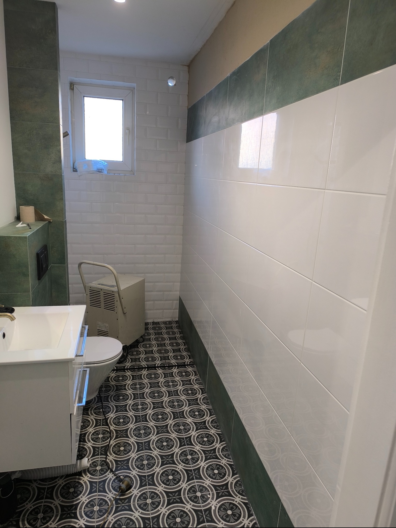 Wykończona łazienka z biało-zielonymi płytkami na ścianach, wzorzystą podłogą i białą umywalką. Widoczny osuszacz powietrza na kółkach.