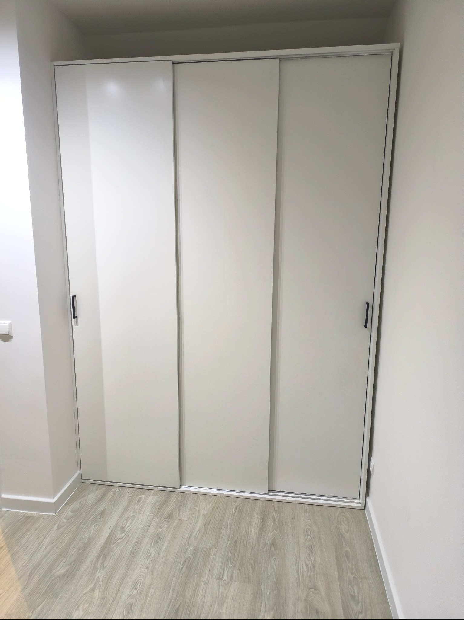 Biała szafa wnękowa z przesuwnymi drzwiami, widoczna na tle jasnych ścian i podłogi imitującej drewno, z minimalistycznymi, pionowymi uchwytami.