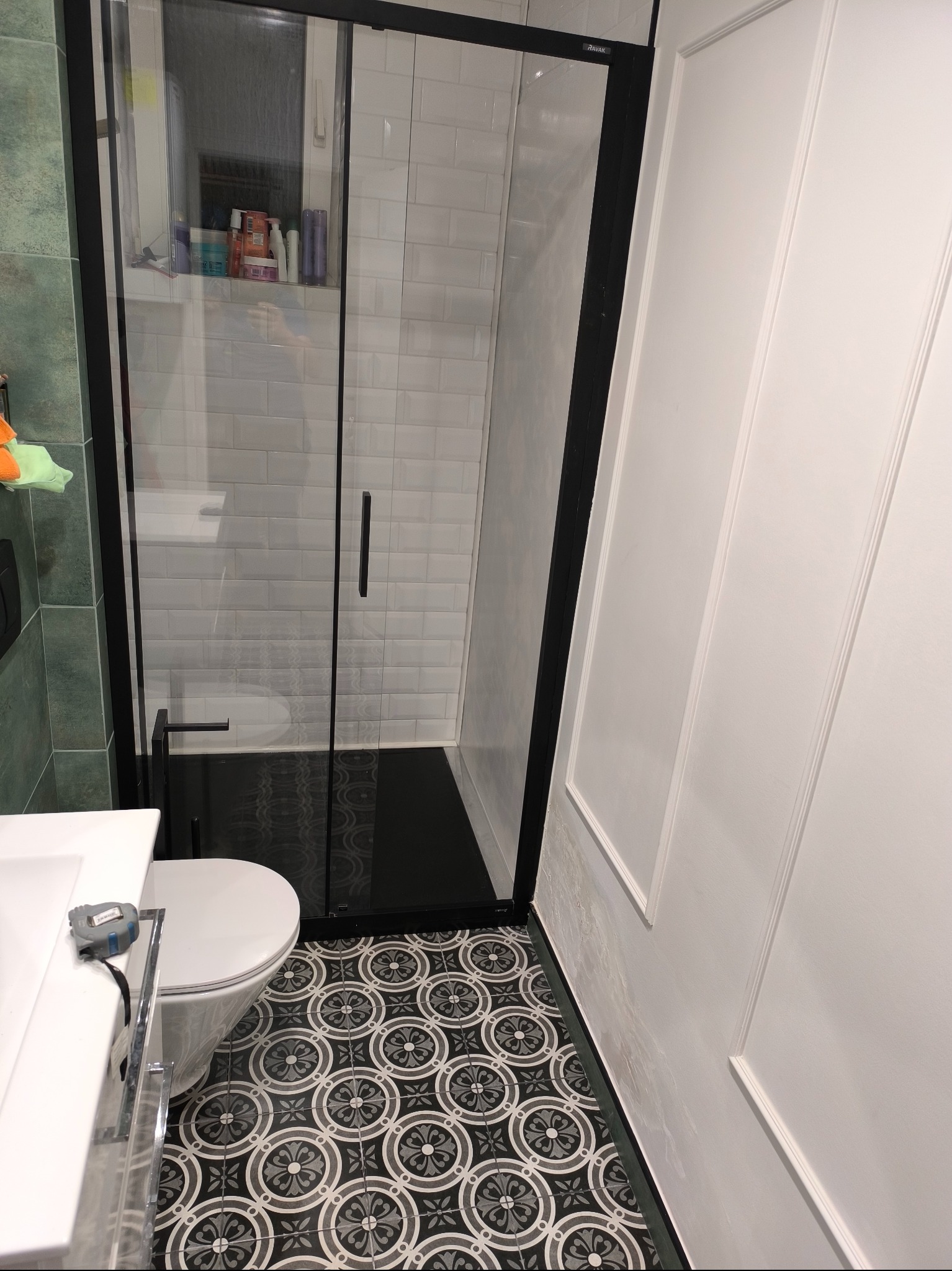 Nowoczesna kabina prysznicowa z czarnymi profilami i brodzikiem, widok na białe płytki na ścianach i dekoracyjną podłogę w geometryczne wzory, fragment toalety i umywalki z widoczną miarką.