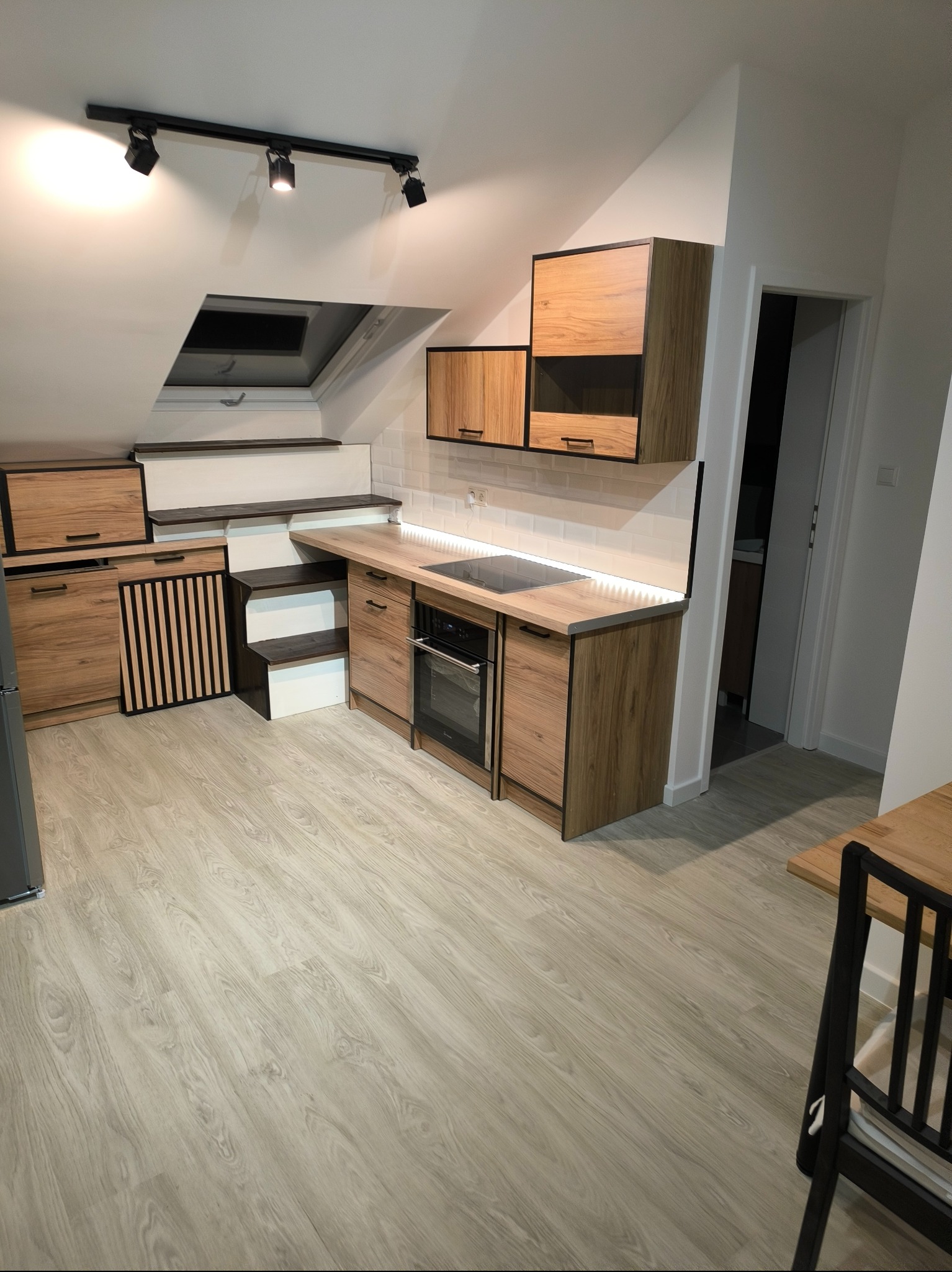 Kuchnia w skosach na poddaszu z drewnianymi szafkami, płytą indukcyjną i piekarnikiem, oświetleniem podszafkowym LED i schodami do okna dachowego