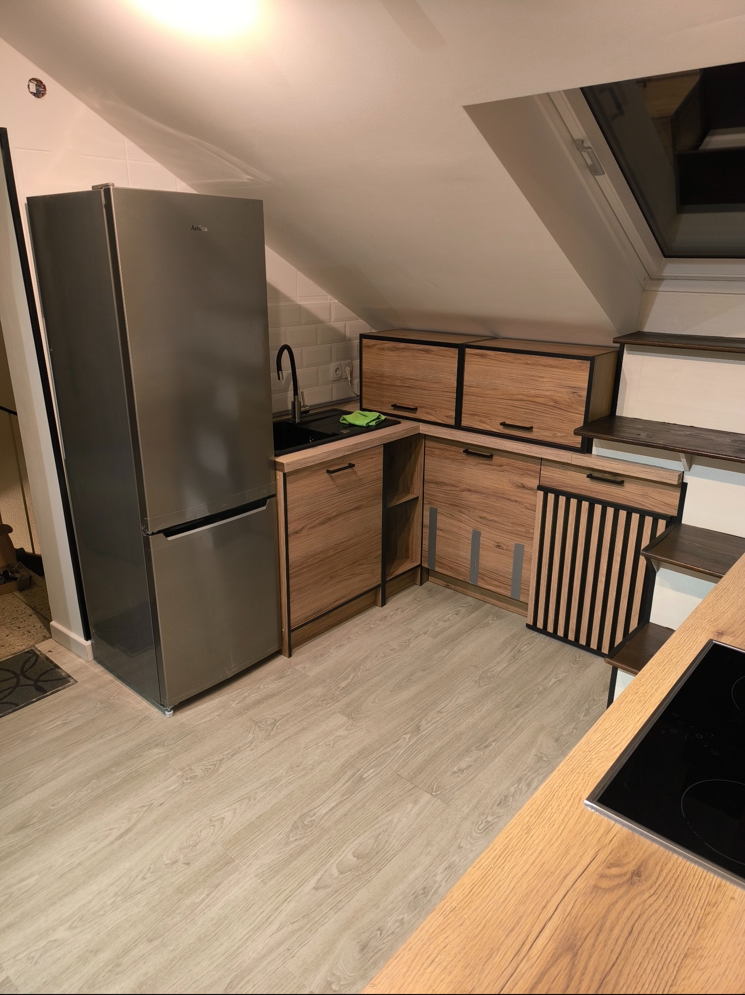 Kuchnia w skosach z zabudową meblową z drewnopodobnych frontów, srebrna lodówka Amica, czarny zlew i płyta indukcyjna, jasna podłoga.