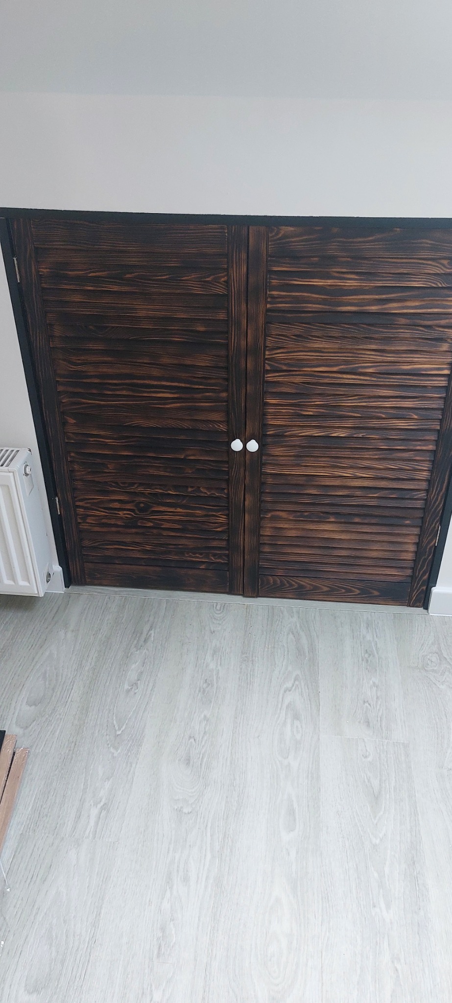 Dwuskrzydłowe, drewniane drzwi przesuwne z żaluzjami, bejcowane na ciemny brąz, z białymi gałkami, zamontowane w białej ścianie, widok z góry na podłogę z paneli.