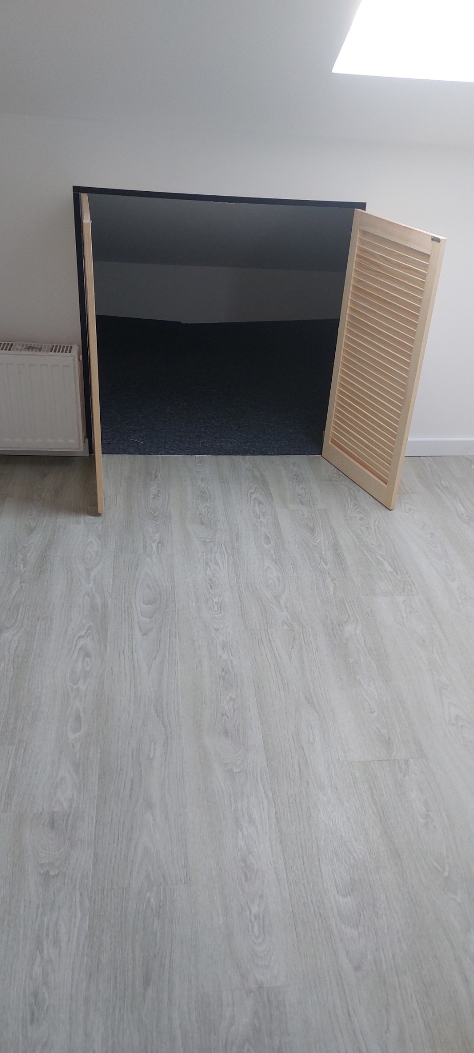 Wejście do schowka pod skosem dachu z drewnianymi, ażurowymi drzwiczkami, widok na szarą wykładzinę i białe ściany, podłoga wyłożona panelami imitującymi drewno.