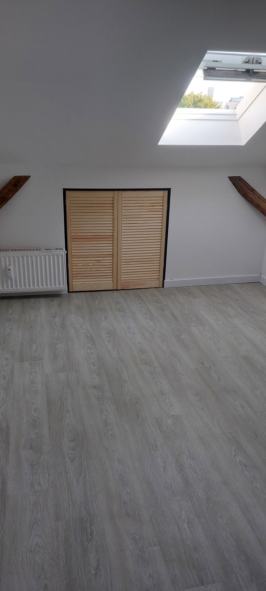 Poddasze z drewnianymi belkami, jasną podłogą, grzejnikiem żeberkowym i zabudową szafy z żaluzjowymi drzwiami na tle okna dachowego.