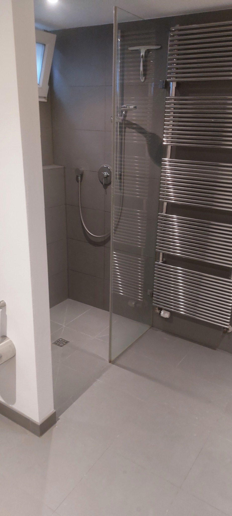 Nowoczesna łazienka z szarą glazurą, kabiną prysznicową ze szklaną ścianką, chromowaną armaturą i grzejnikiem drabinkowym. Widoczny odpływ liniowy w podłodze.