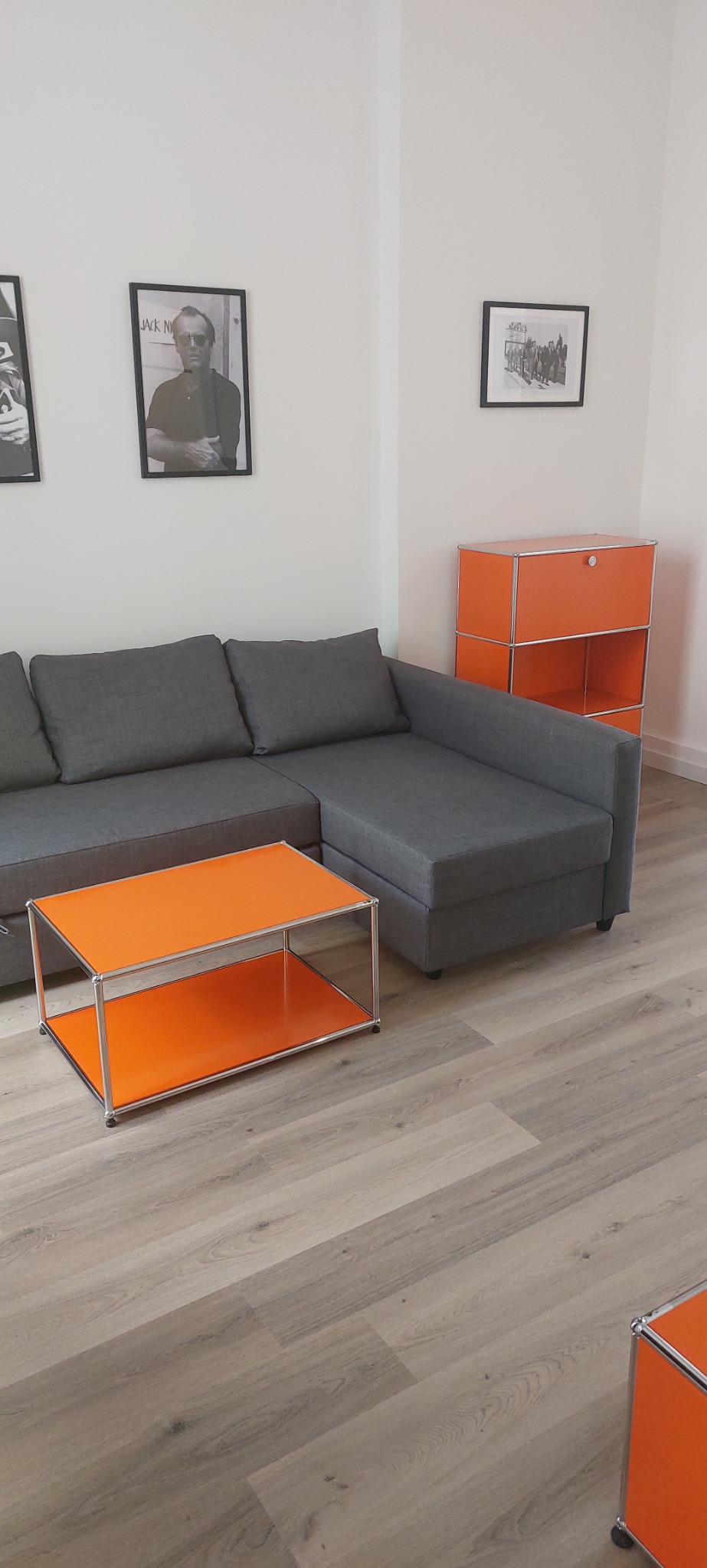 Szary narożnik w salonie z laminowaną podłogą, pomarańczowy stolik kawowy i szafka modułowa w tym samym kolorze, czarno-białe zdjęcia w ramkach na ścianie.
