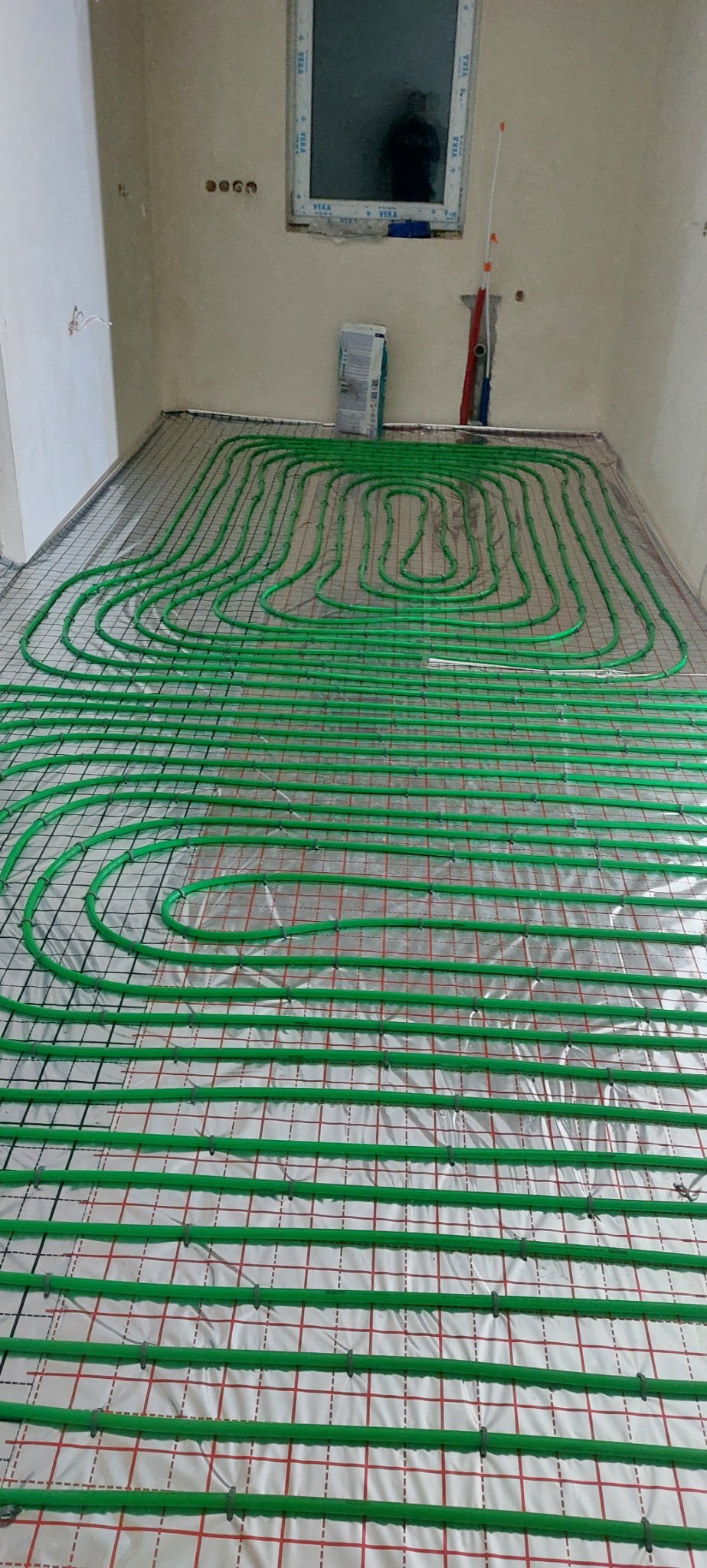 Rozłożona instalacja ogrzewania podłogowego z zielonych rurek na folii z siatką, widoczne okno i ściany w trakcie wykańczania.