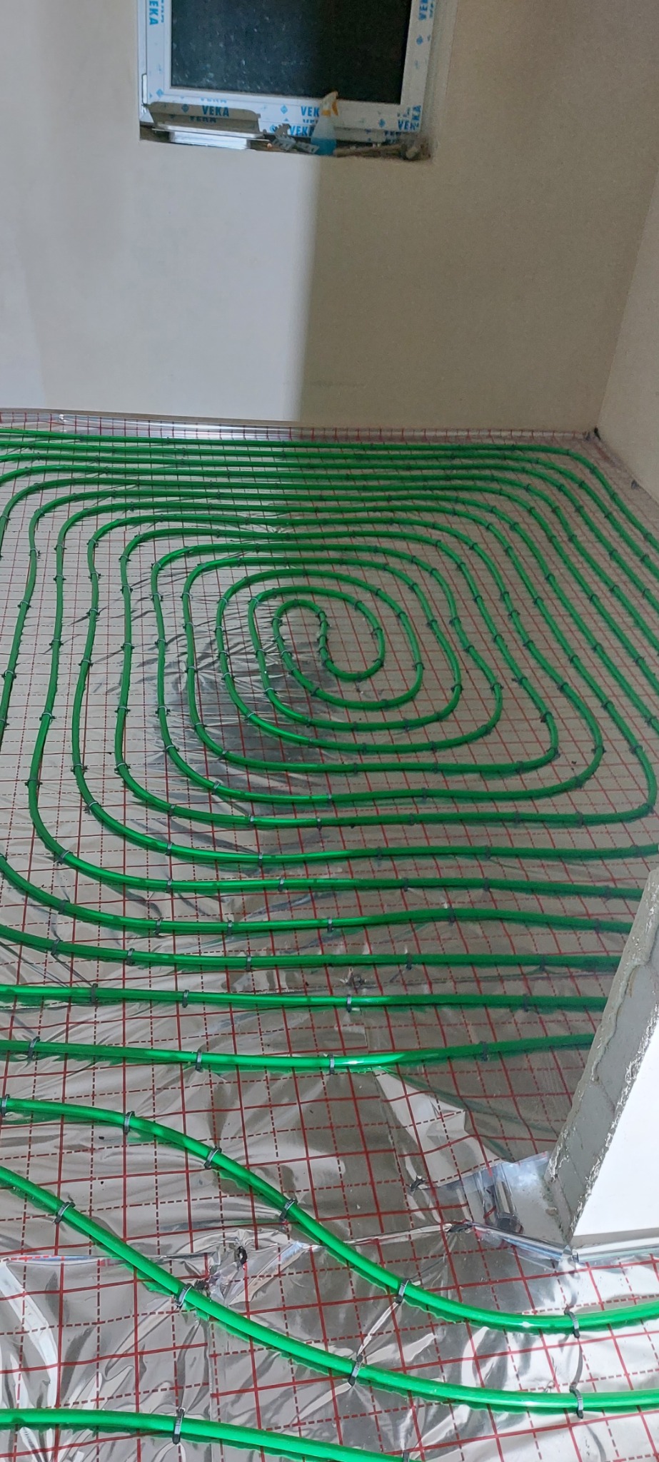 Zielone rurki ogrzewania podłogowego ułożone spiralnie na folii izolacyjnej z czerwoną siatką, widok z góry na etapie montażu.