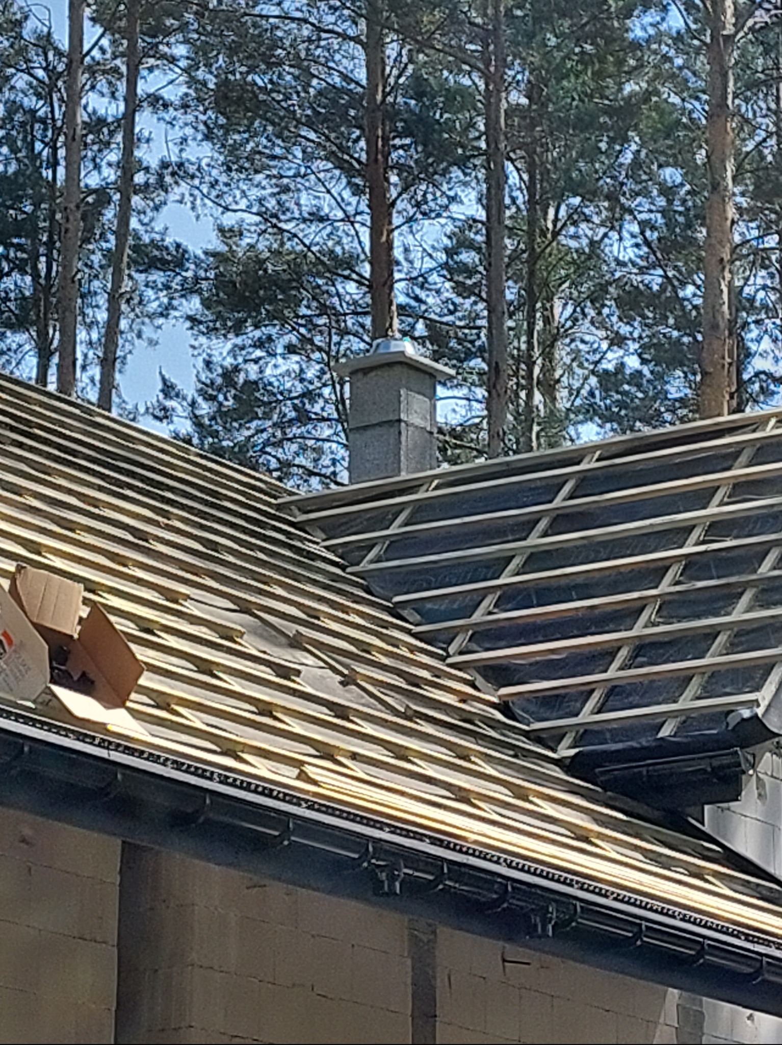 Konstrukcja więźby dachowej z łatami i kontrłatami, widoczne czarne membrany, komin z szarej cegły, rynna i fragment ściany budynku w tle drzewa.