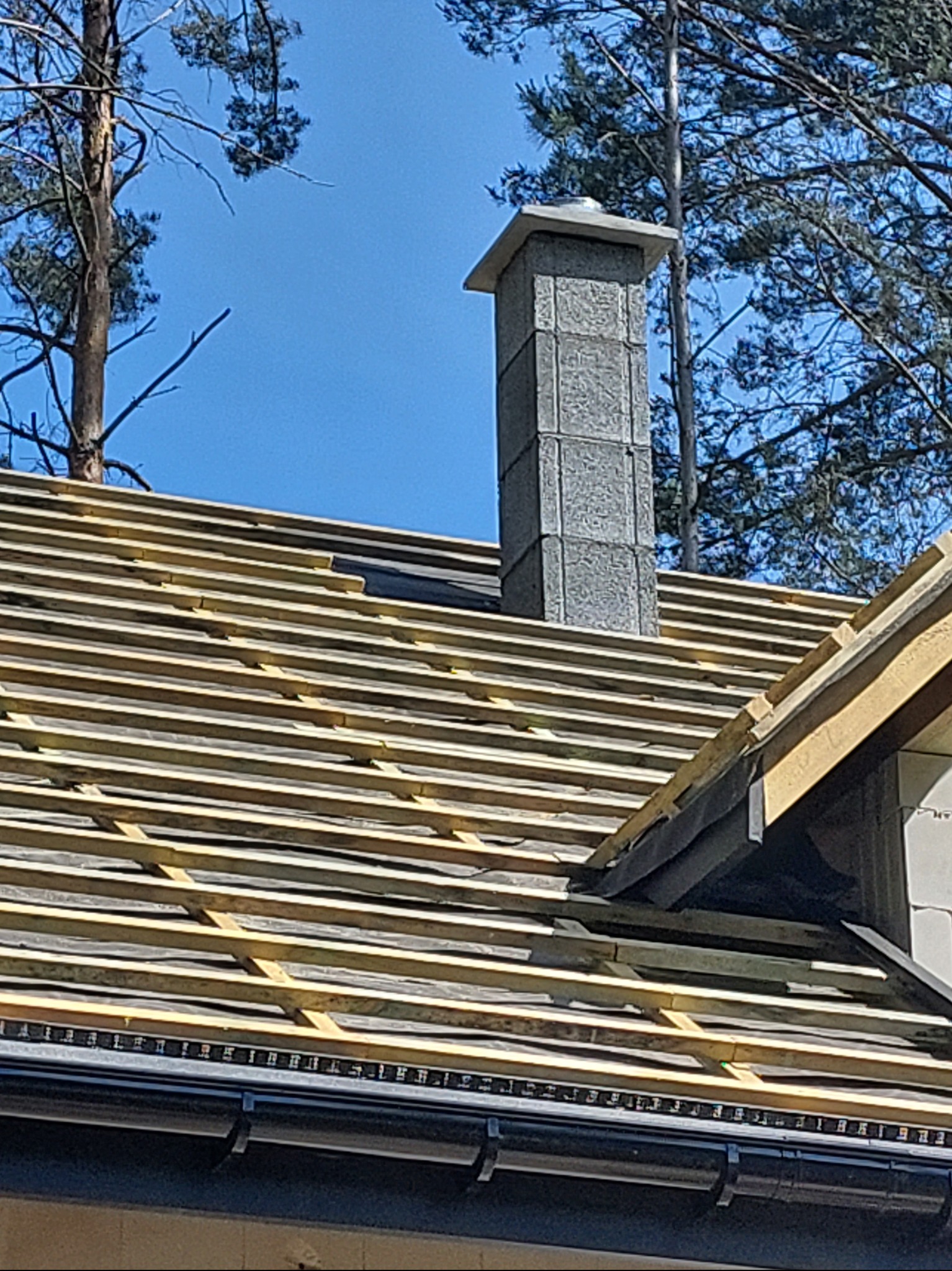 Surowy stan konstrukcji dachu z widocznym kominem z szarego kamienia na tle błękitnego nieba i drzew. Na dachu widoczne drewniane łaty i czarna membrana dachowa. Komin z betonowych bloczków.