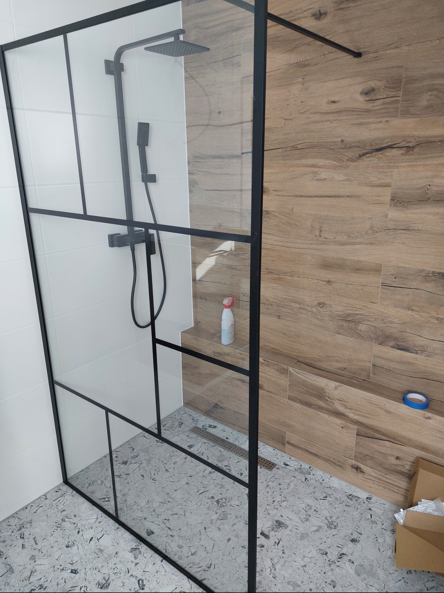 Nowoczesna kabina prysznicowa z czarną ramą, szklaną ścianką działową, deszczownicą i słuchawką prysznicową na tle białych płytek i drewnopodobnych paneli, widoczny odpływ liniowy w posadzce...