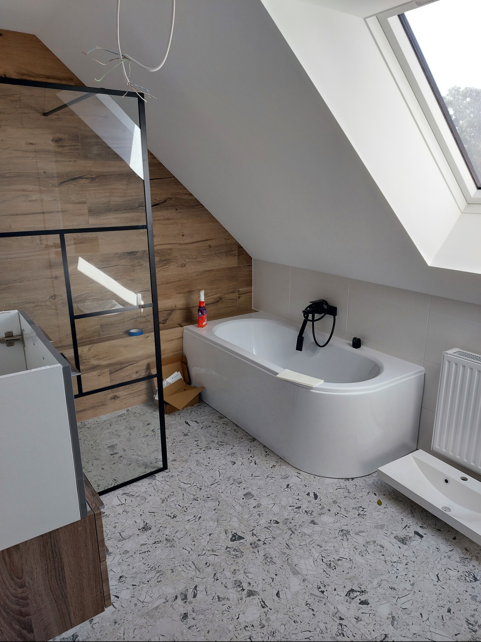 Wnętrze łazienki na poddaszu w Sękocinie Nowy w trakcie remontu, z widoczną wanną, kabiną prysznicową z czarnymi profilami, umywalką i grzejnikiem. Uwagę zwracają drewnopodobne płytki na ścianie...