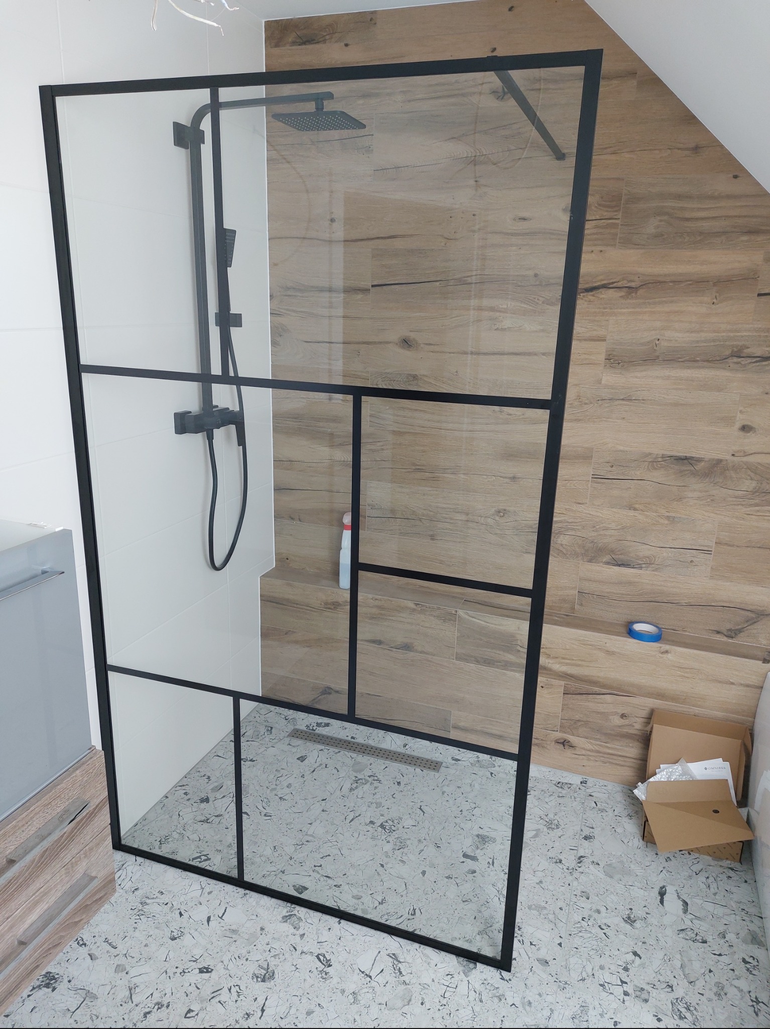 Nowoczesna kabina prysznicowa z czarnymi profilami na tle białych i drewnopodobnych płytek, widoczny odpływ liniowy i armatura w czarnym macie.