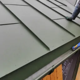 Prace dekarskie: montaż paneli dachowych w kolorze oliwkowej zieleni, widoczna rynna z elementami mocującymi oraz fragment komina zabezpieczonego folią na tle zalesionego wzgórza.