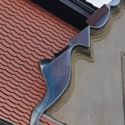 Fragment dachu z ceramiczną dachówką karpiówką i ozdobnym, metalowym zakończeniem rynny przy elewacji budynku.