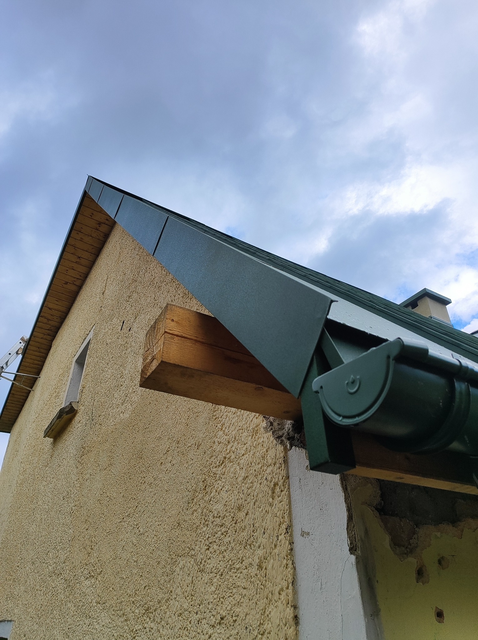 Fragment dachu z nowym, ciemnozielonym orynnowaniem, widoczne mocowanie rynny do drewnianej belki wystającej ze ściany budynku o żółtej, chropowatej elewacji, na tle pochmurnego nieba.