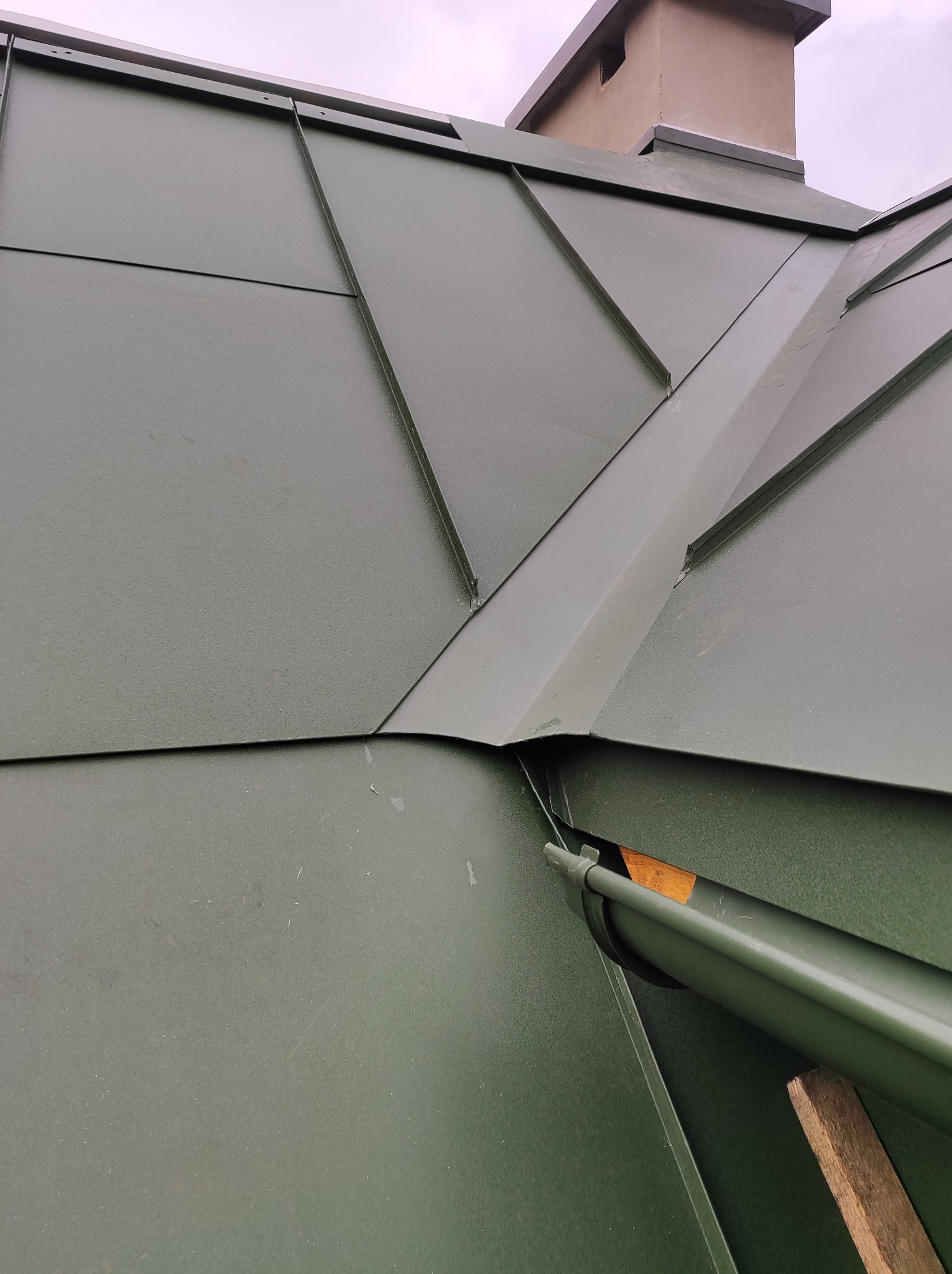 Szczegółowe ujęcie fragmentu dachu z blachy w odcieniach zieleni, z widocznymi obróbkami blacharskimi i fragmentem rynny, uchwycone pod kątem.