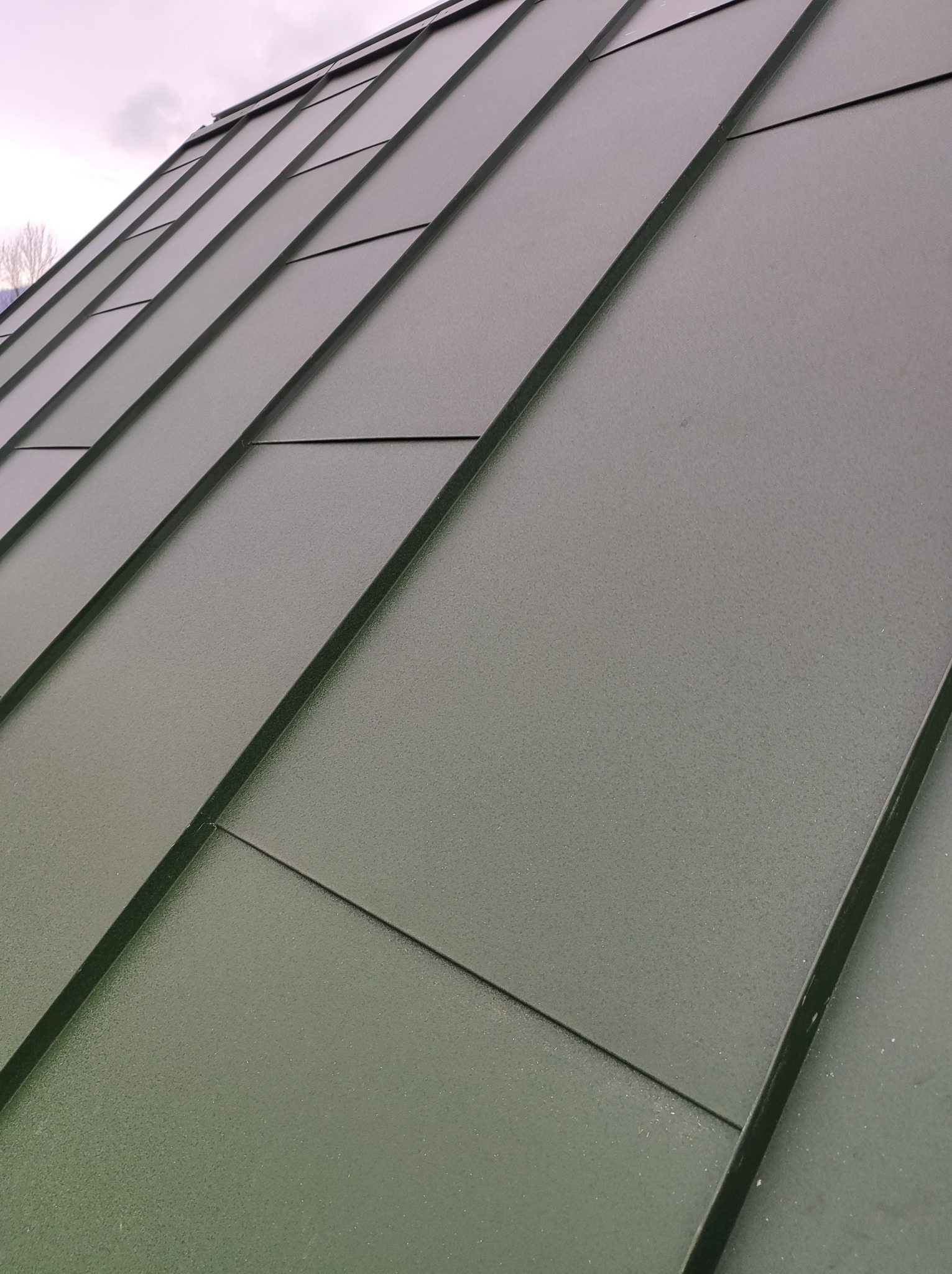 Szczegółowe ujęcie ciemnozielonego dachu z blachy na rąbek stojący, widoczne połączenia paneli i faktura materiału, sfotografowane pod kątem.