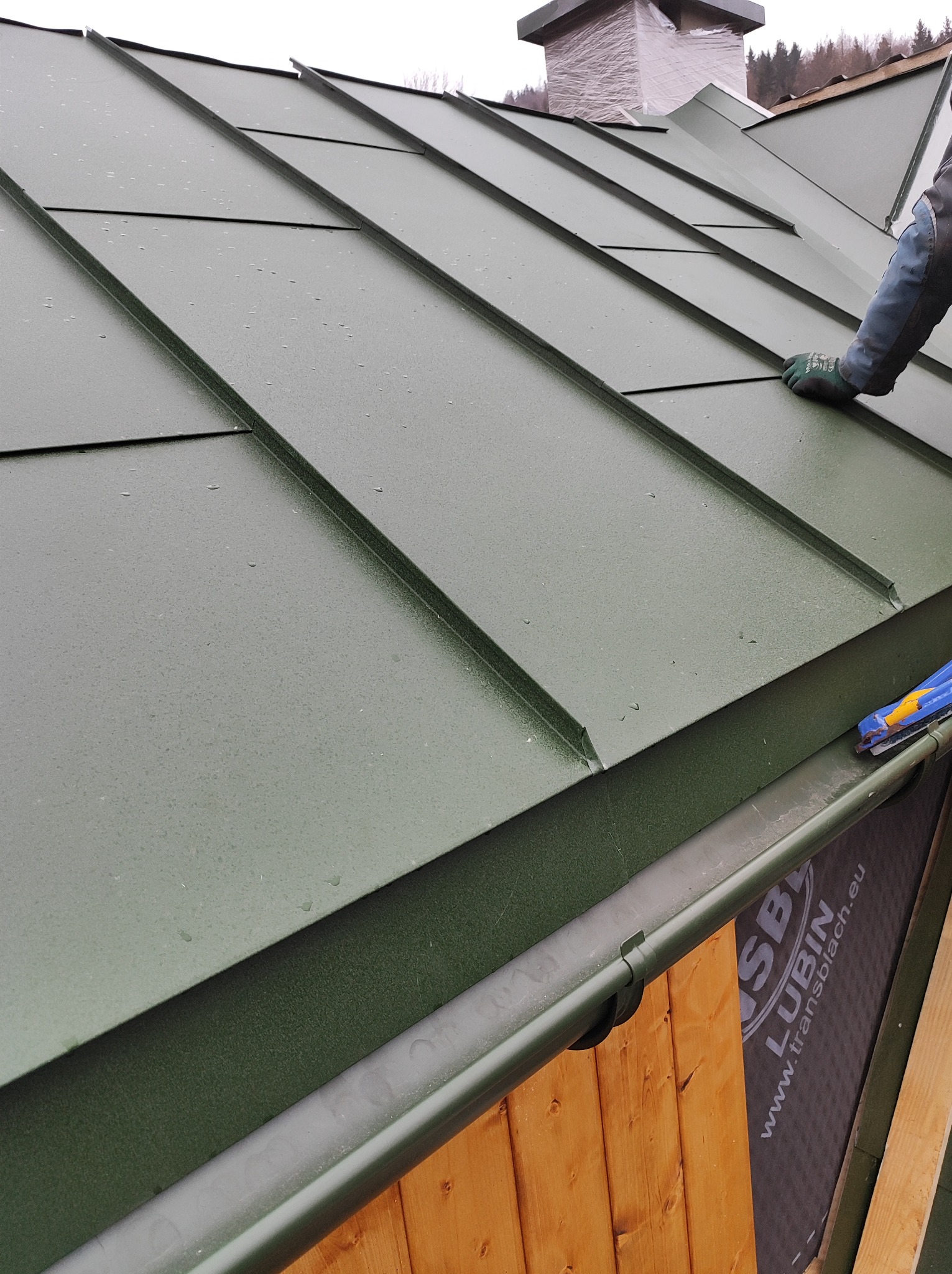 Prace dekarskie: montaż paneli dachowych w kolorze oliwkowej zieleni, widoczna rynna z elementami mocującymi oraz fragment komina zabezpieczonego folią na tle zalesionego wzgórza.