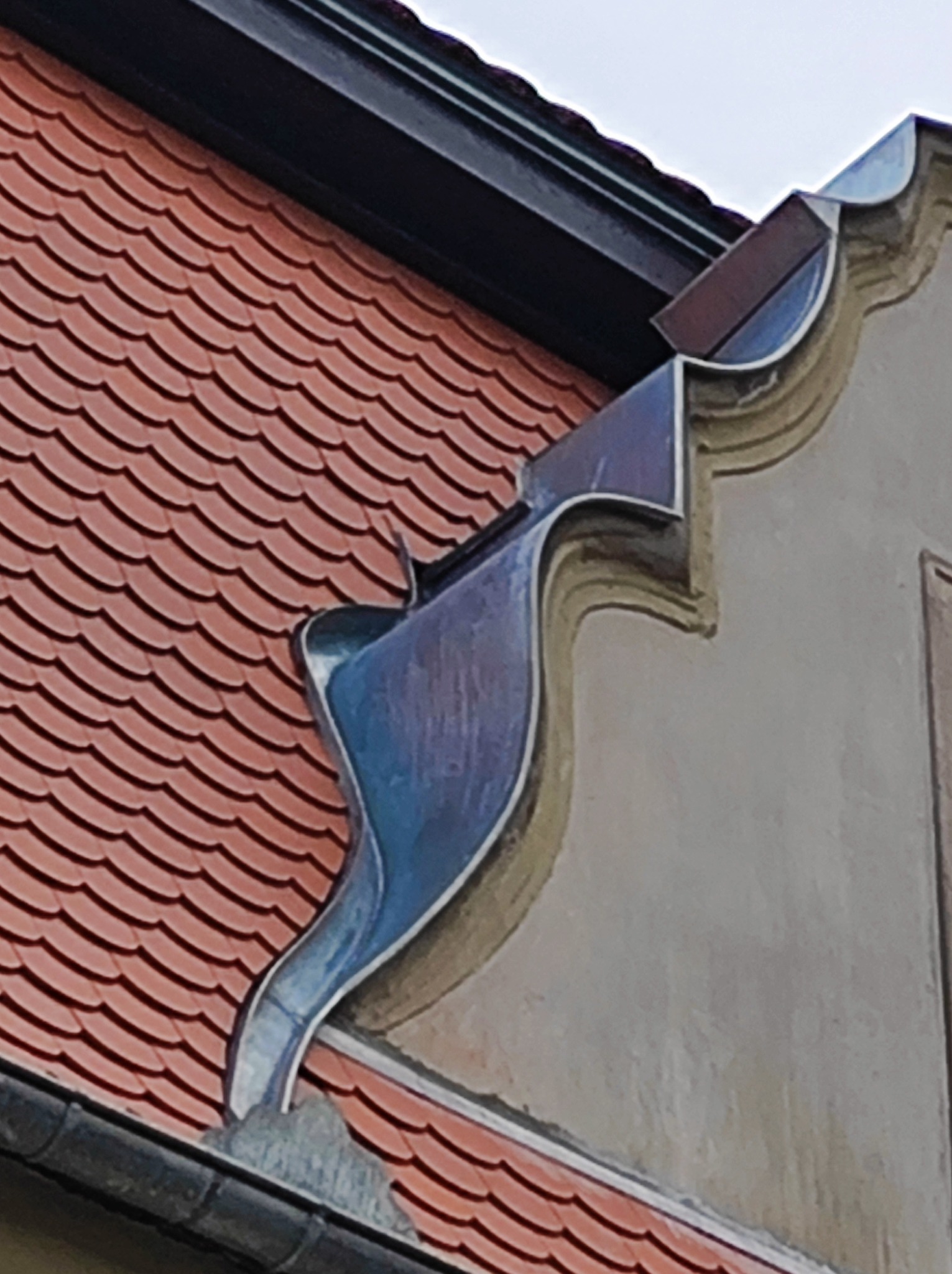 Fragment dachu z ceramiczną dachówką karpiówką i ozdobnym, metalowym zakończeniem rynny przy elewacji budynku.