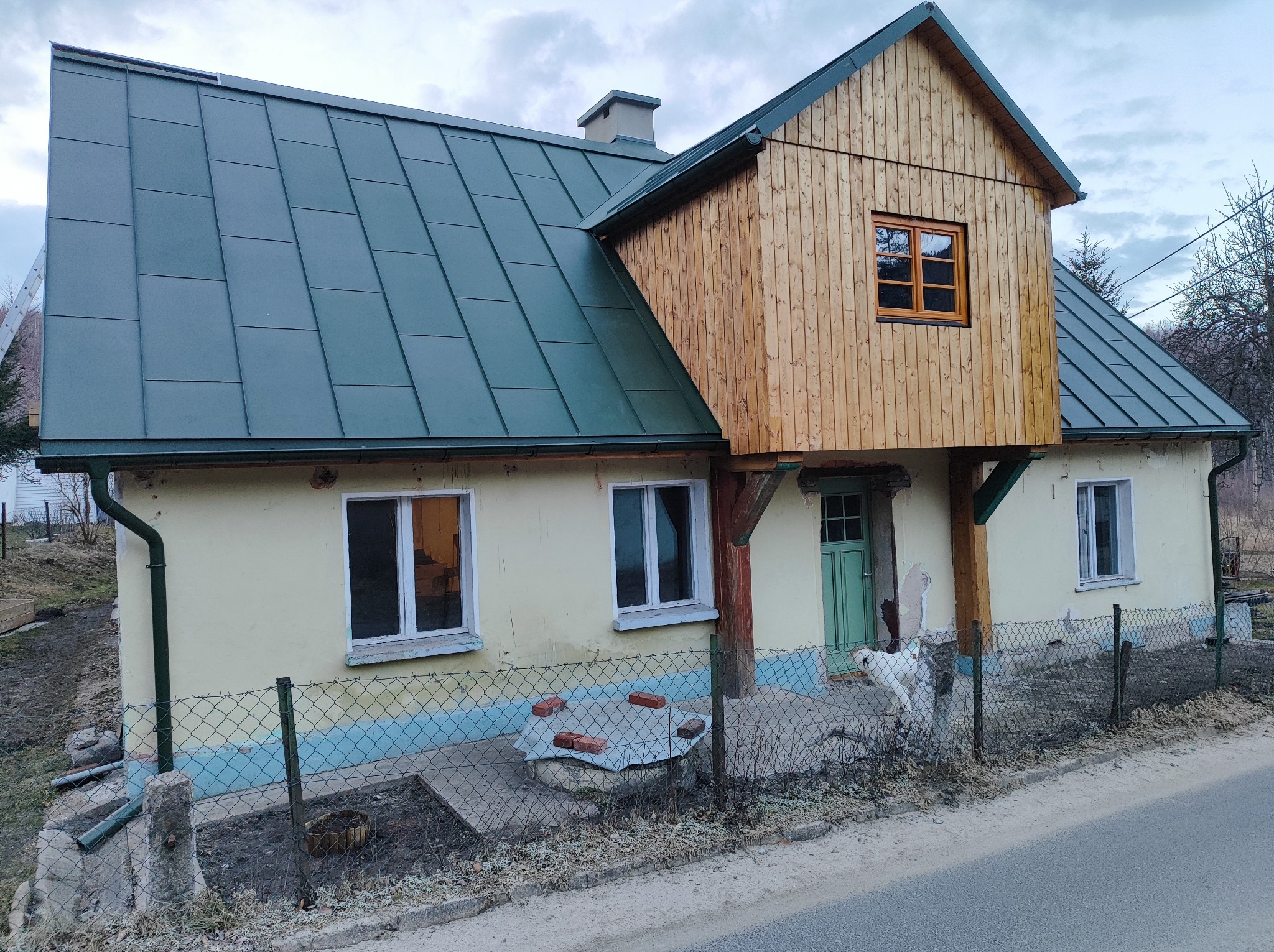 Dom z nowym, ciemnozielonym dachem z blachy trapezowej i drewnianą nadbudówką z oknem, widoczne rynny, elewacja częściowo odnowiona, biały pies przy ogrodzeniu.