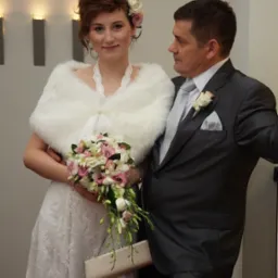 Para młoda: panna młoda w białej sukni z koronkowym wzorem, futrzanym bolerku i bukietem różowych i białych kwiatów, obok pan młody w ciemnoszarym garniturze z butonierką z białej róży.