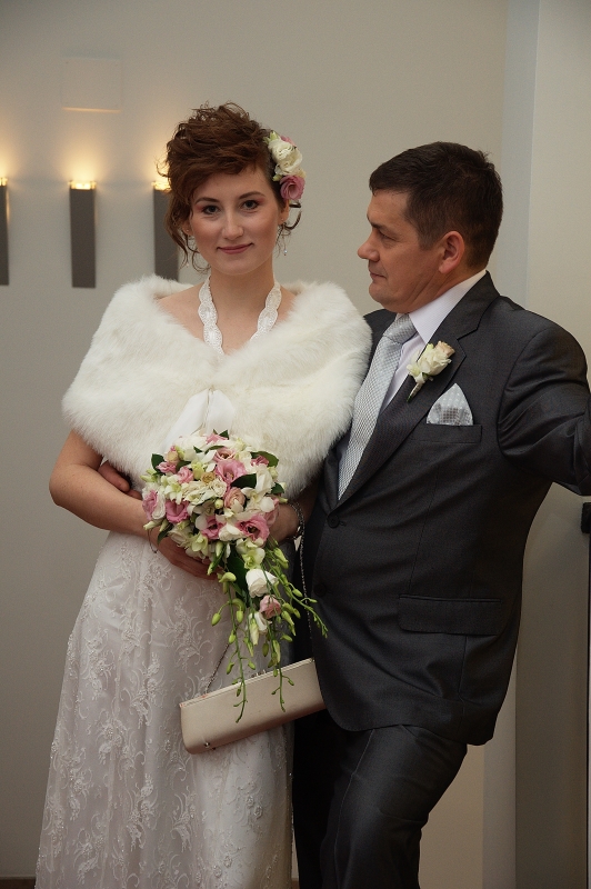 Para młoda: panna młoda w białej sukni z koronkowym wzorem, futrzanym bolerku i bukietem różowych i białych kwiatów, obok pan młody w ciemnoszarym garniturze z butonierką z białej róży.