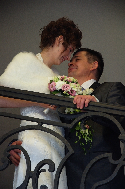 Uśmiechnięta para młoda, panna młoda w białym futerku i koronkowej sukni trzyma bukiet różowych i białych kwiatów, opierając się o czarną, ozdobną balustradę.