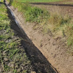 Długi, wykopany rów melioracyjny z niewielką ilością wody na dnie, biegnący wzdłuż pola uprawnego w wiejskim krajobrazie.