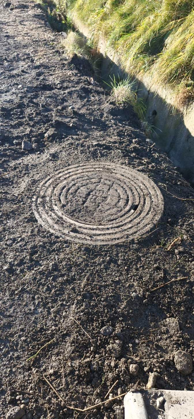 Okragła, betonowa pokrywa studzienki kanalizacyjnej, częściowo przysypana ziemią, z widocznym betonowym korytkiem odpływowym i trawą w tle.
