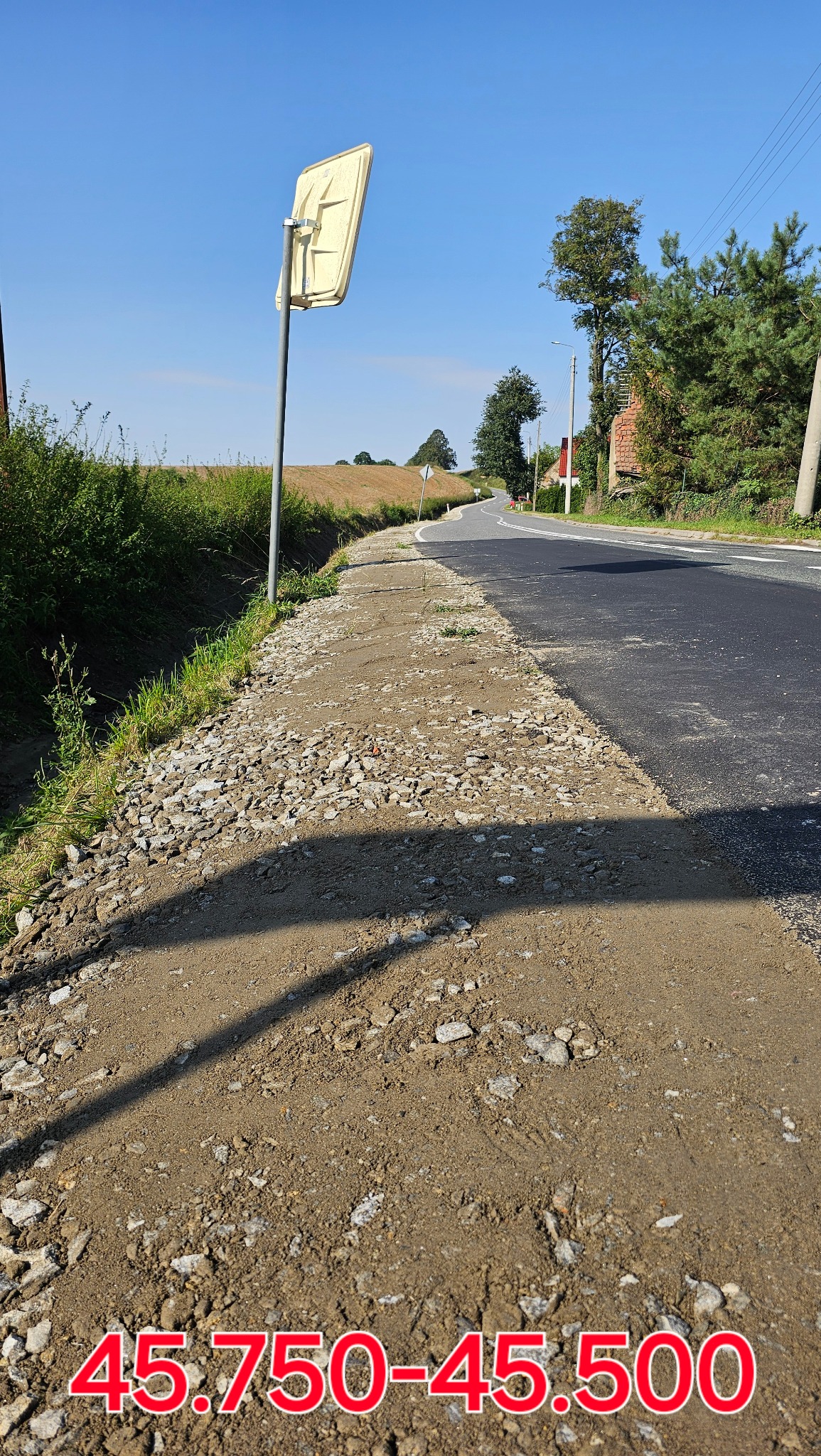 Fragment drogi z nowo utwardzonym poboczem, widoczne kruszywo i ziemia, obok asfaltowa nawierzchnia i znak drogowy.