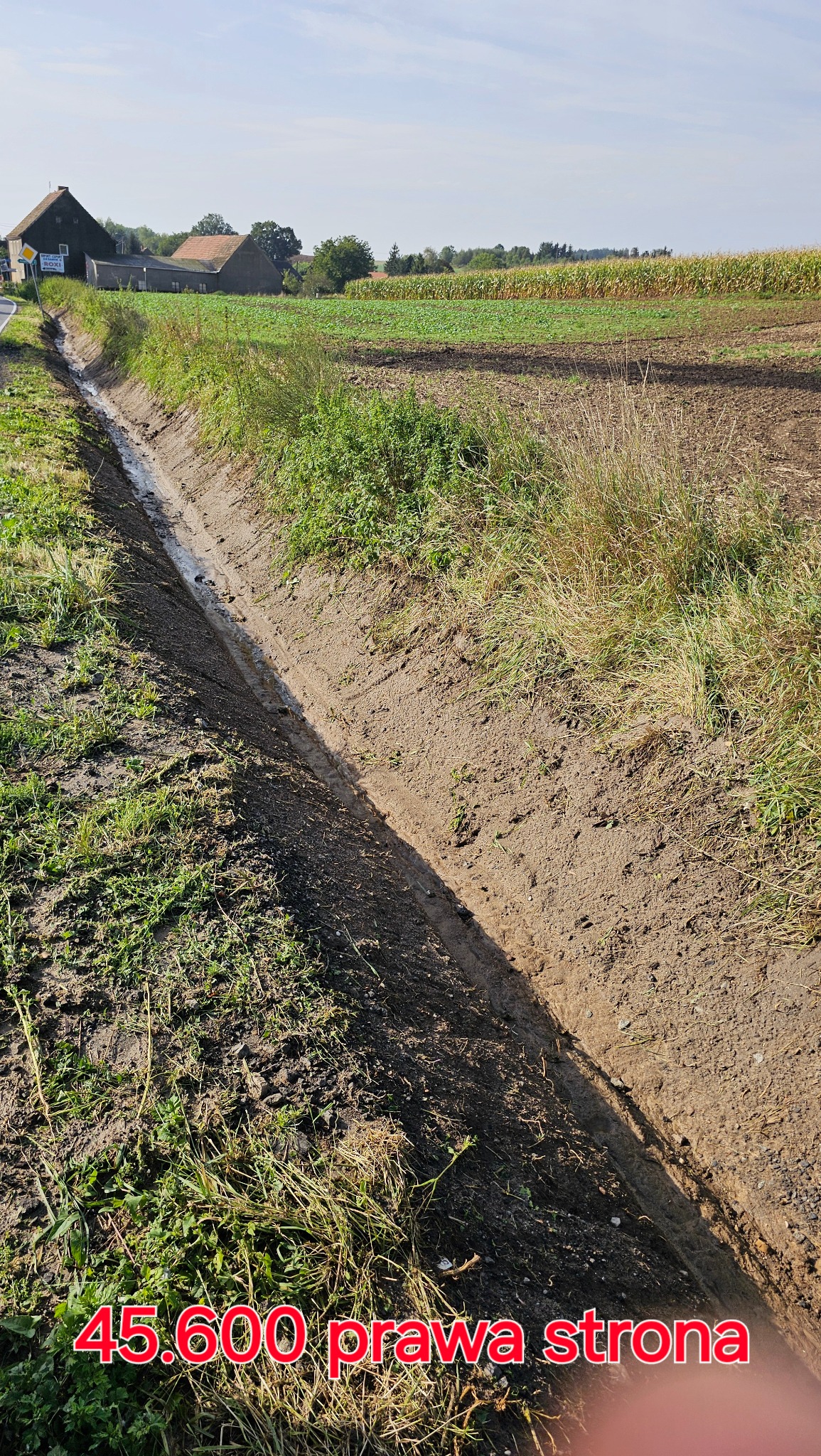 Długi, wykopany rów melioracyjny z niewielką ilością wody na dnie, biegnący wzdłuż pola uprawnego w wiejskim krajobrazie.