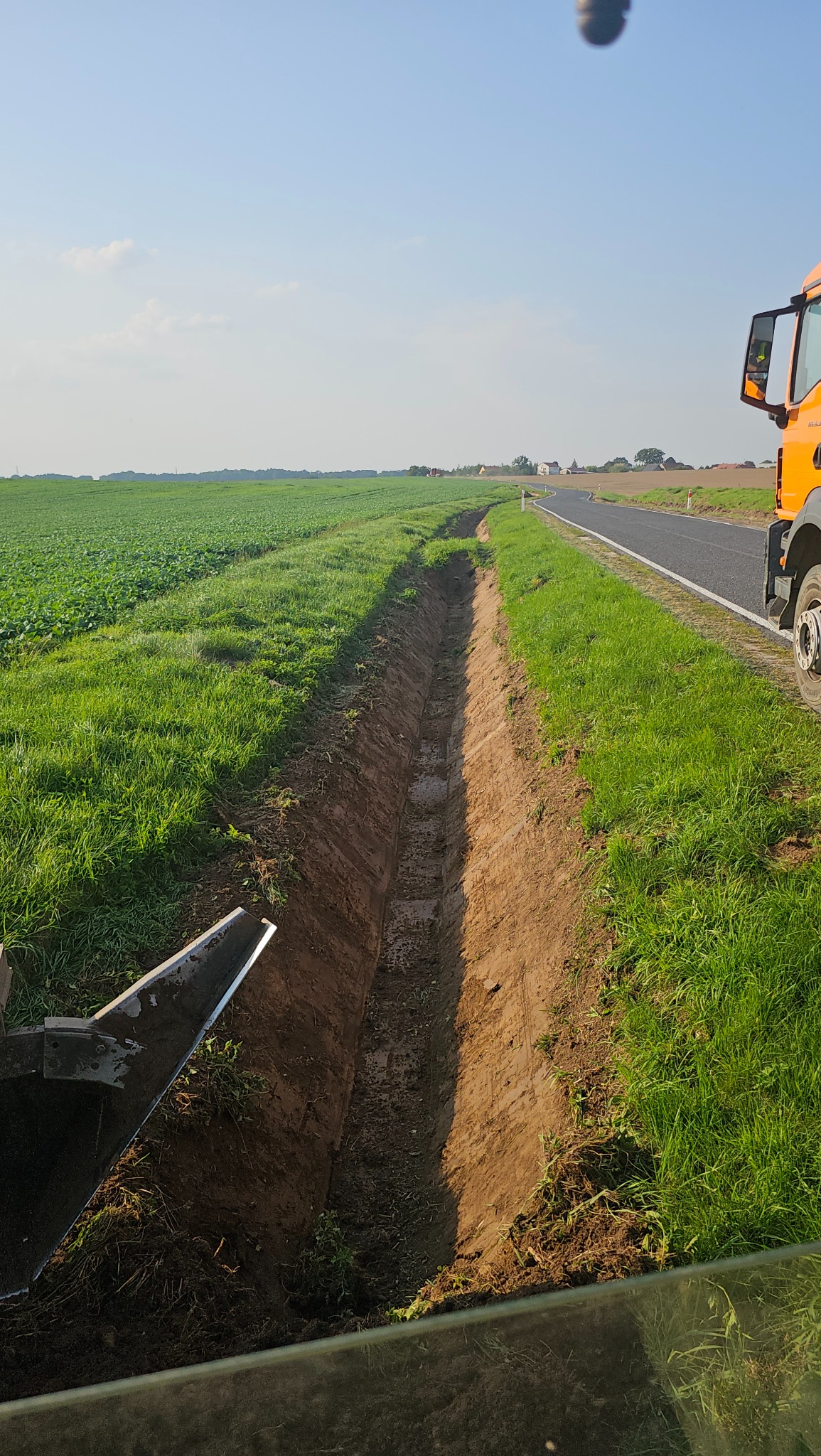 Wykopany rów w ziemi wzdłuż drogi, widoczny fragment pomarańczowej ciężarówki i pole w tle.