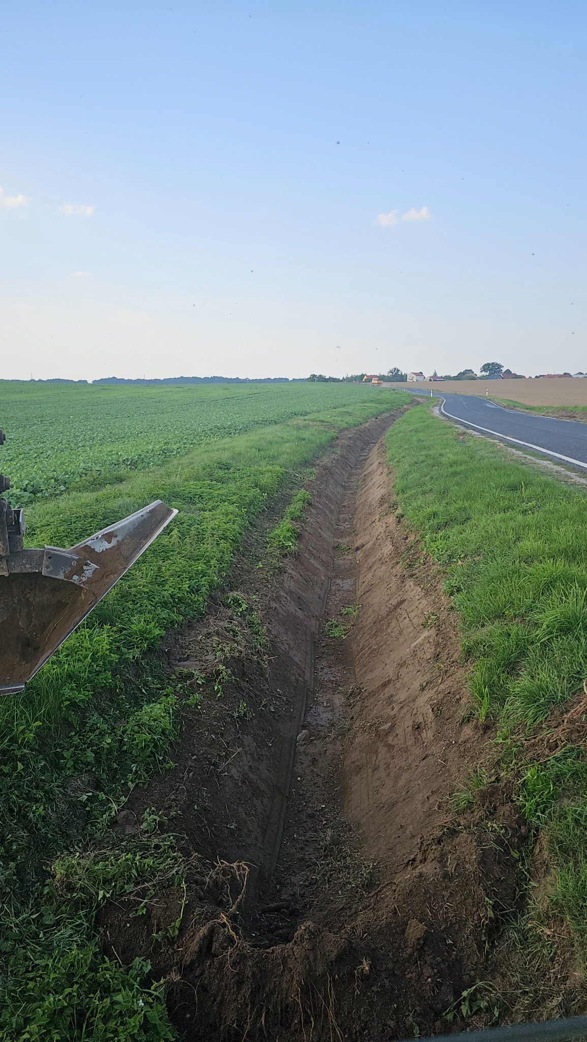 Wykopany rów melioracyjny wzdłuż pola uprawnego i asfaltowej drogi, widoczny fragment łyżki koparki.