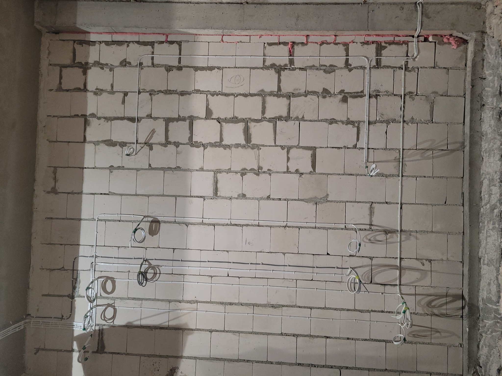 Białe rurki instalacji elektrycznej ułożone na surowej ścianie z betonu komórkowego, widoczne kable i przewody. Etap prac instalacyjnych w budynku.