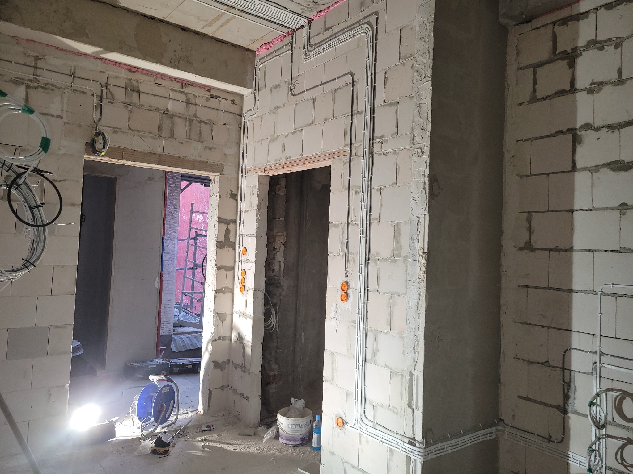 Instalacja elektryczna w surowym wnętrzu: białe kable prowadzone po ścianach z bloczków, widoczne puszki instalacyjne, lampa budowlana oświetla przestrzeń.