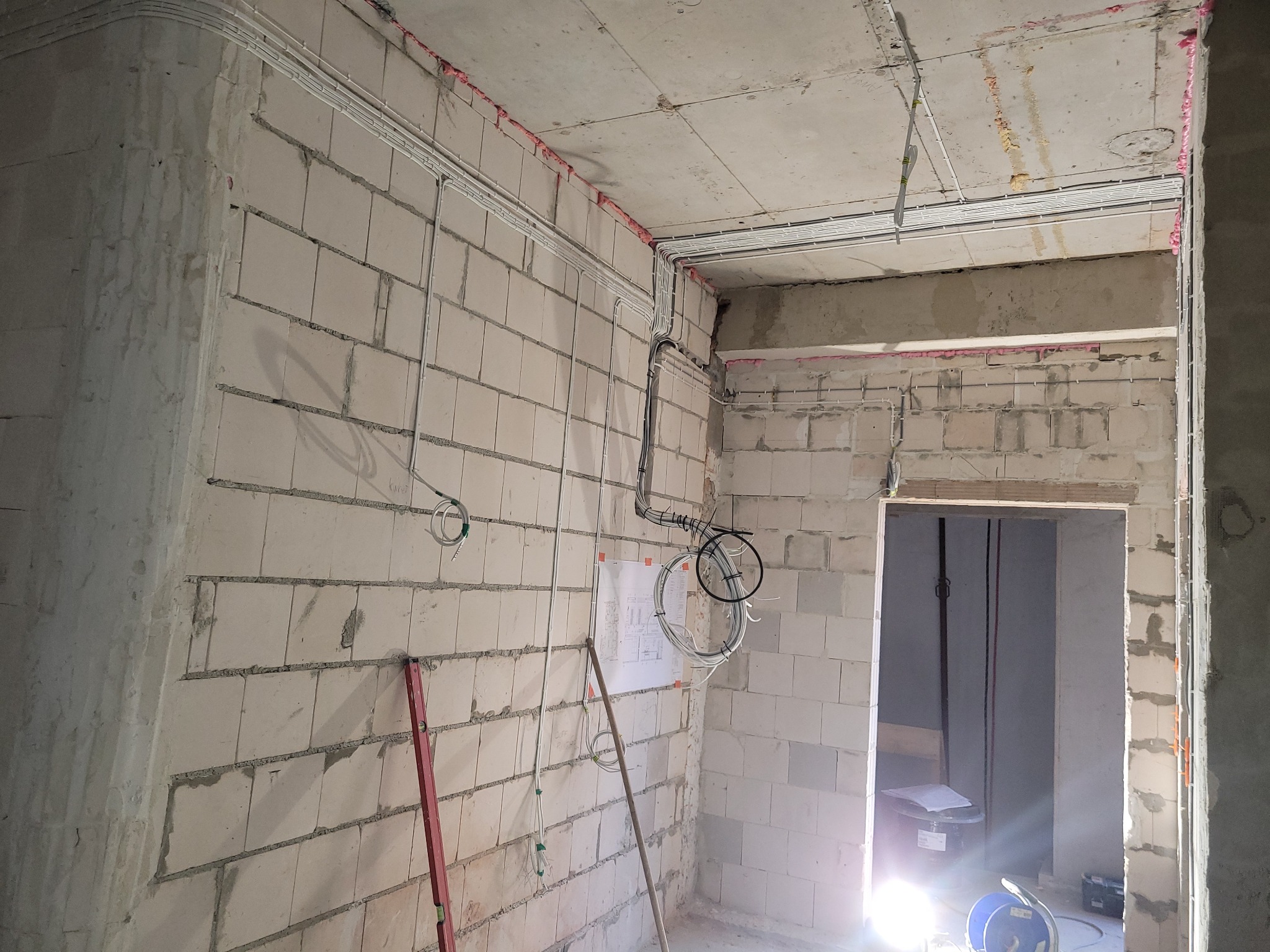 Wnętrze w trakcie prac elektrycznych: widoczne kable w peszlach ułożone na ścianach z betonu komórkowego, poziomica oparta o ścianę, plan instalacji na ścianie.