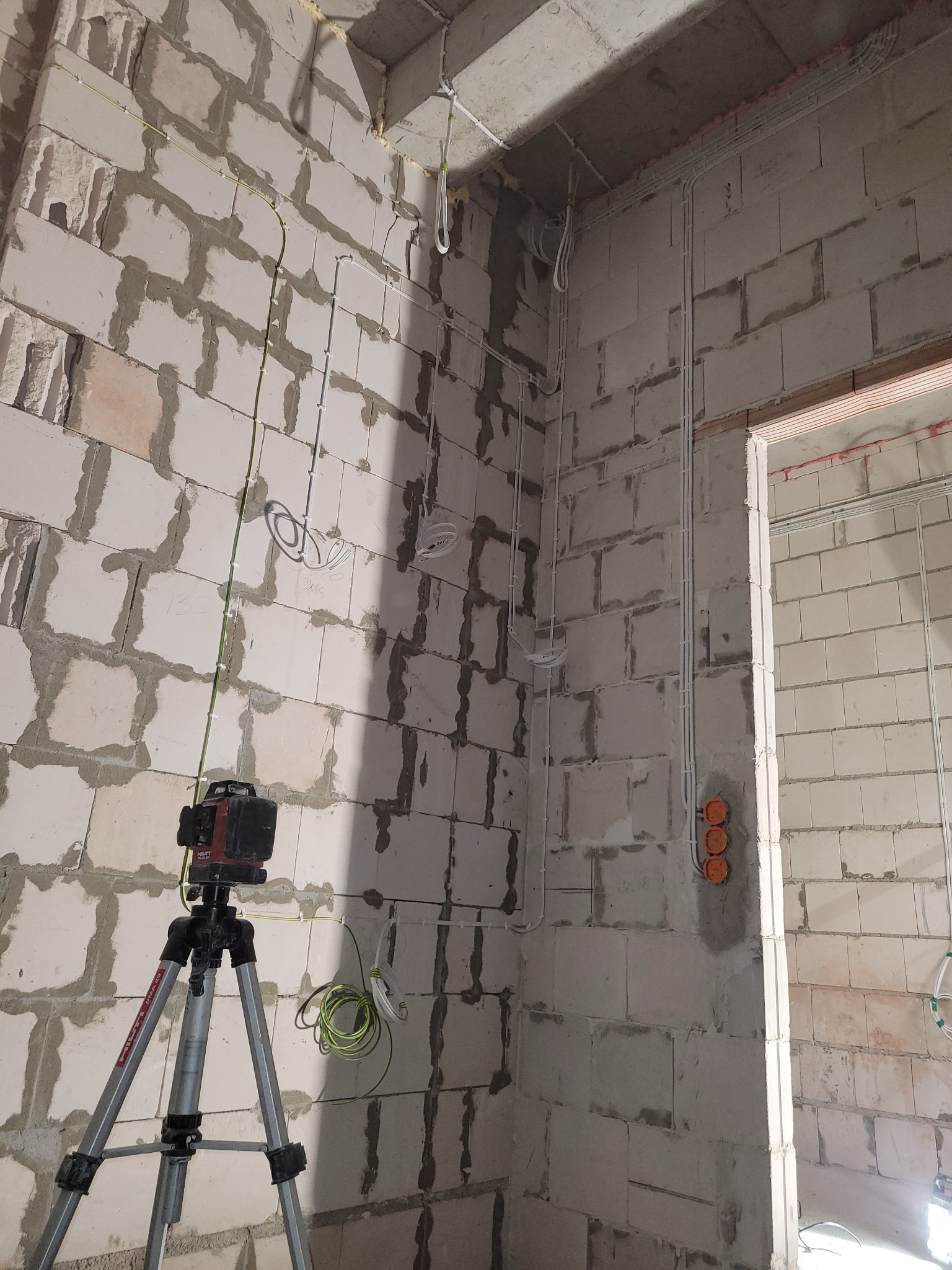 Wnętrze w trakcie prac elektrycznych: widoczne kable, puszki instalacyjne i laser krzyżowy na statywie, na tle surowych ścian z betonu komórkowego.