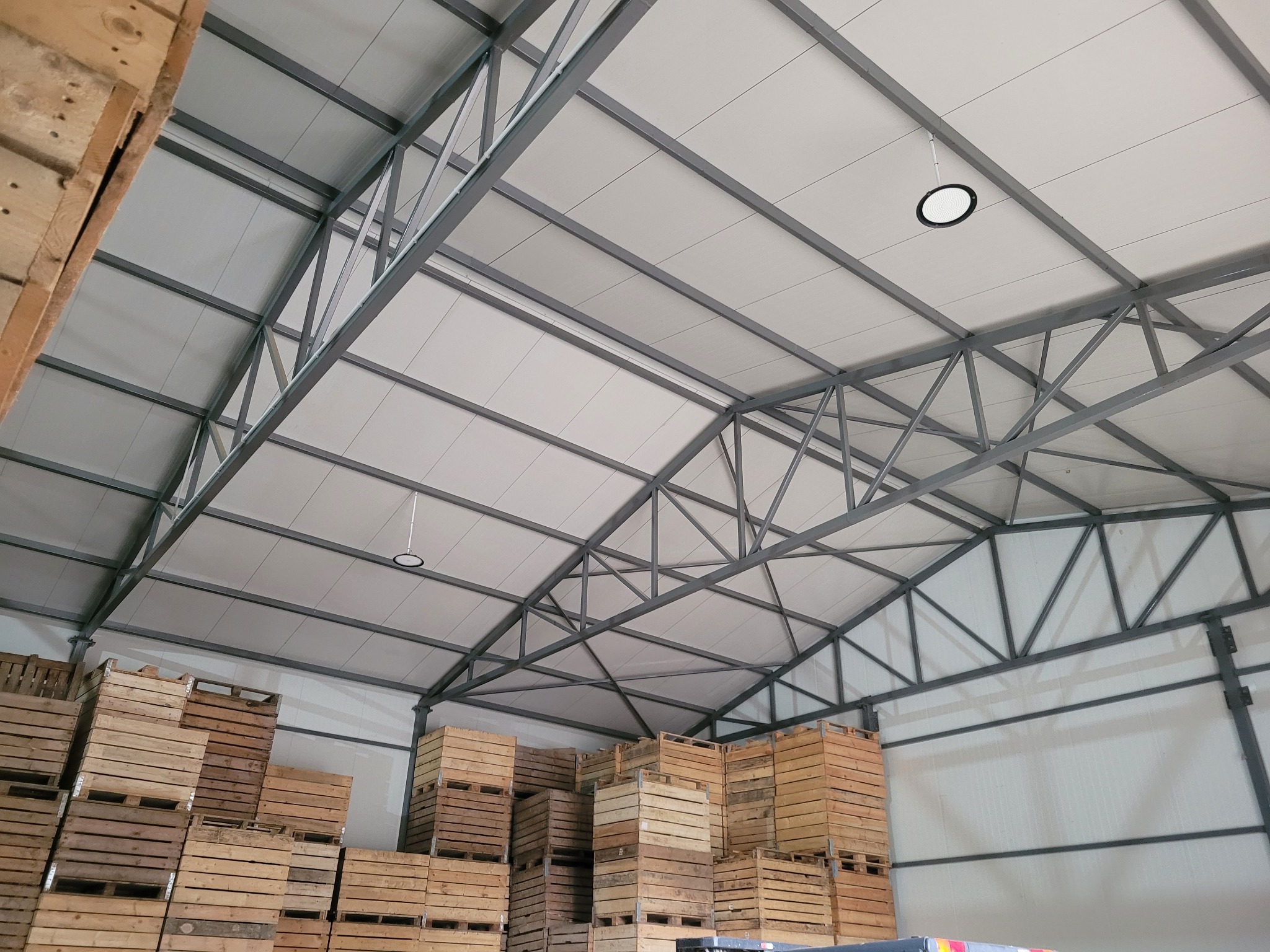 Wnętrze hali magazynowej z metalową konstrukcją dachu i oświetleniem. Ustawione drewniane skrzynie. Widok z dołu, oświetlenie LED.