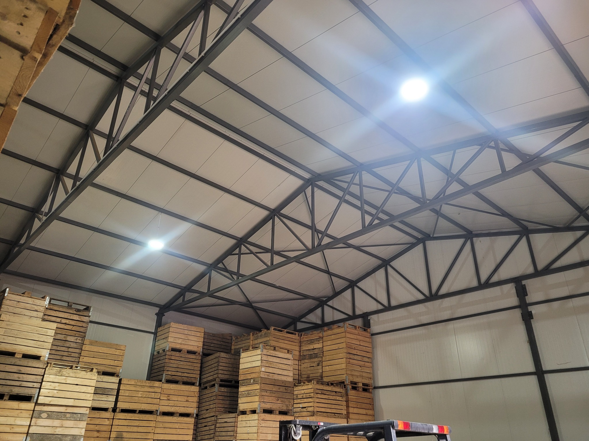 Wnętrze hali magazynowej z metalową konstrukcją dachu i oświetleniem LED, wypełnionej drewnianymi skrzyniami ułożonymi w stosy. Widoczny fragment wózka widłowego.