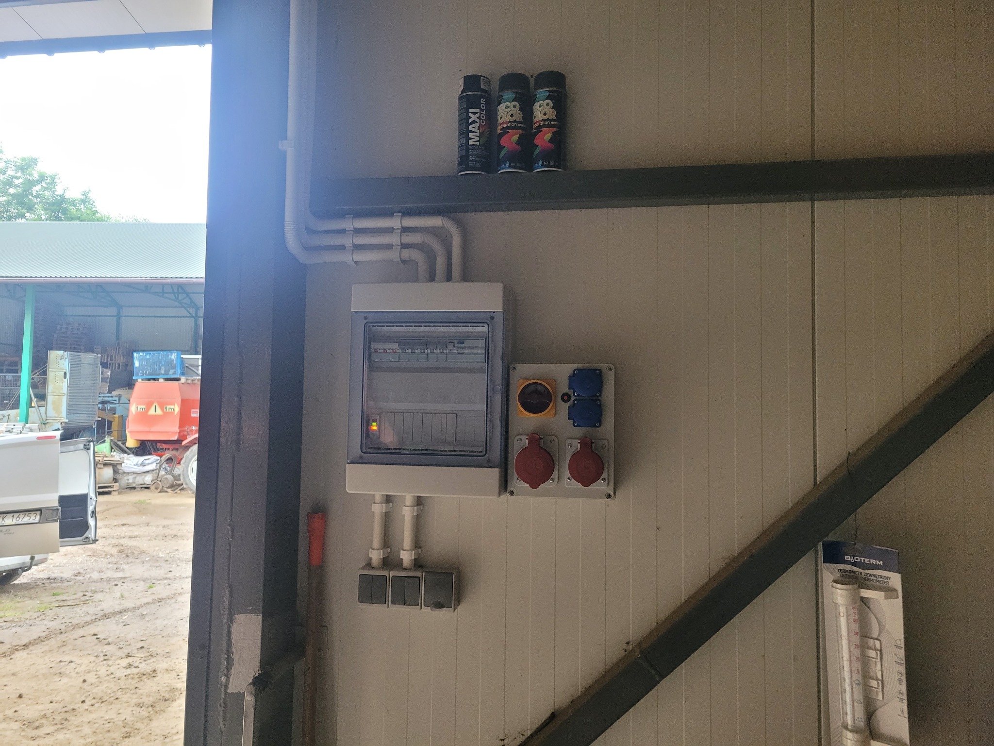 Szara skrzynka elektryczna z bezpiecznikami, gniazdami i włącznikami na panelowej ścianie w hali. Nad nią puszki farb w sprayu. Widoczne kable i rury instalacyjne.