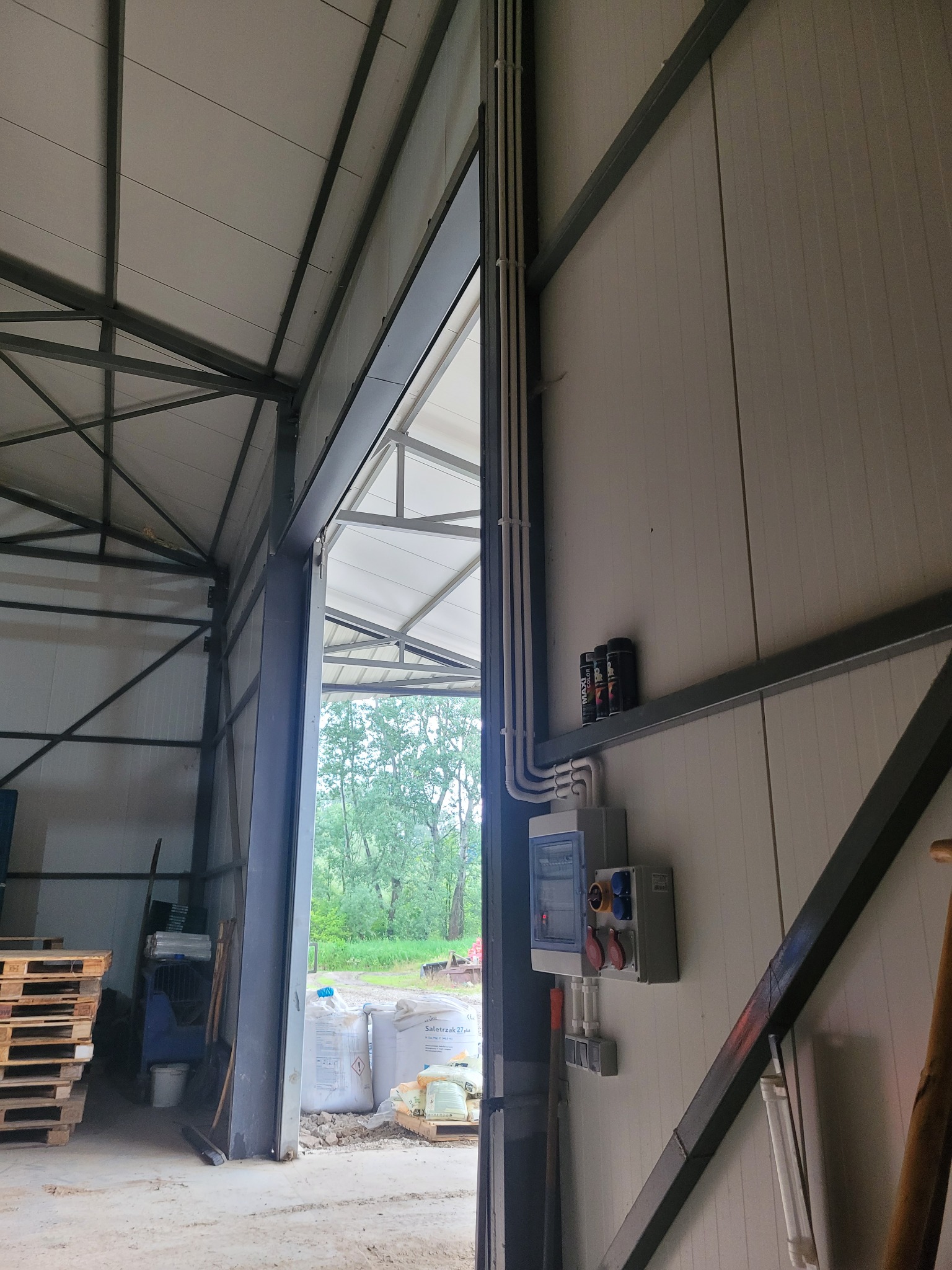 Rozdzielnia elektryczna z przewodami w hali magazynowej. Widoczne zabezpieczenia i gniazda. W tle otwarte wrota i palety.