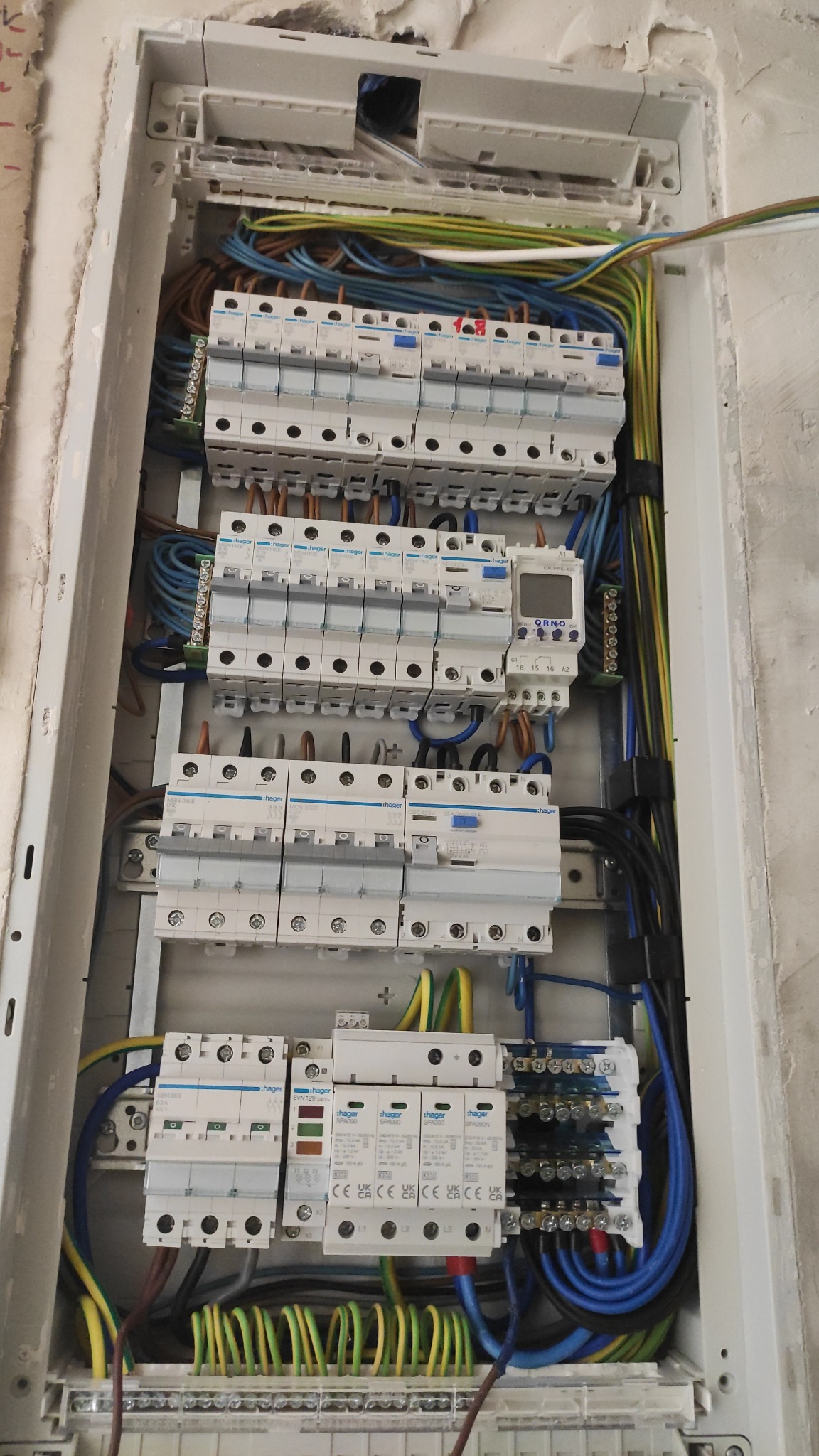 Rozdzielnia elektryczna w obudowie, widoczne zabezpieczenia, przewody w różnych kolorach. Instalacja w trakcie montażu, surowe wykończenie ściany.