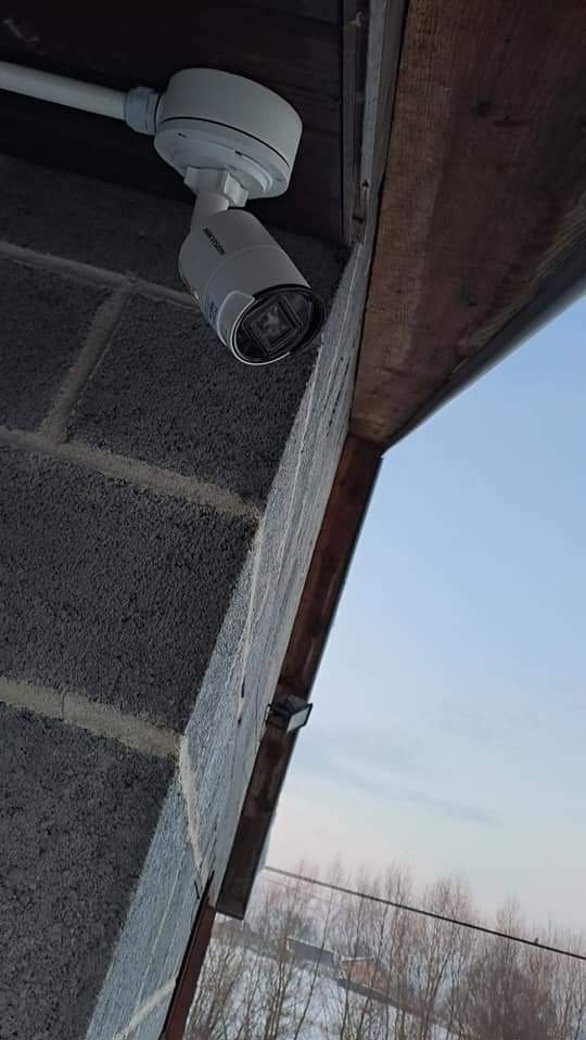 Kamera monitoringu Hikvision zamontowana na szarej ścianie z bloczków, widok z dołu, fragment nieba w tle. Widoczny przewód zasilający.