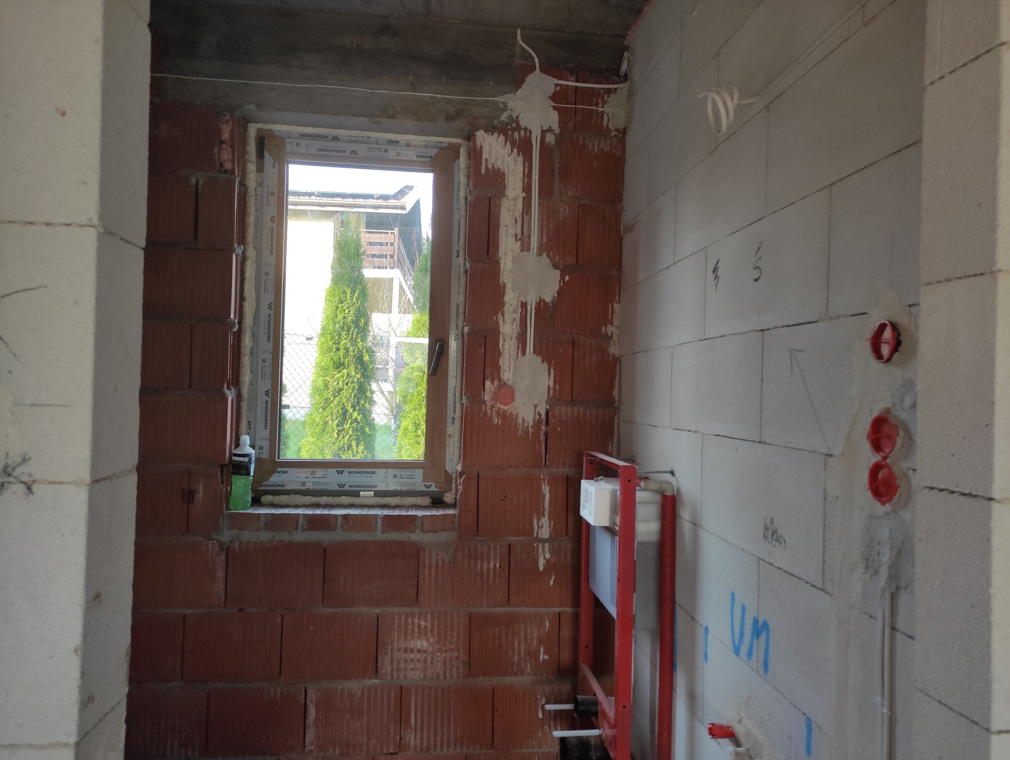 Wnętrze w trakcie prac instalacyjnych: okno z widokiem na zieleń, instalacja elektryczna w ścianie z cegły, fragment instalacji wodnej na ścianie z betonu komórkowego.
