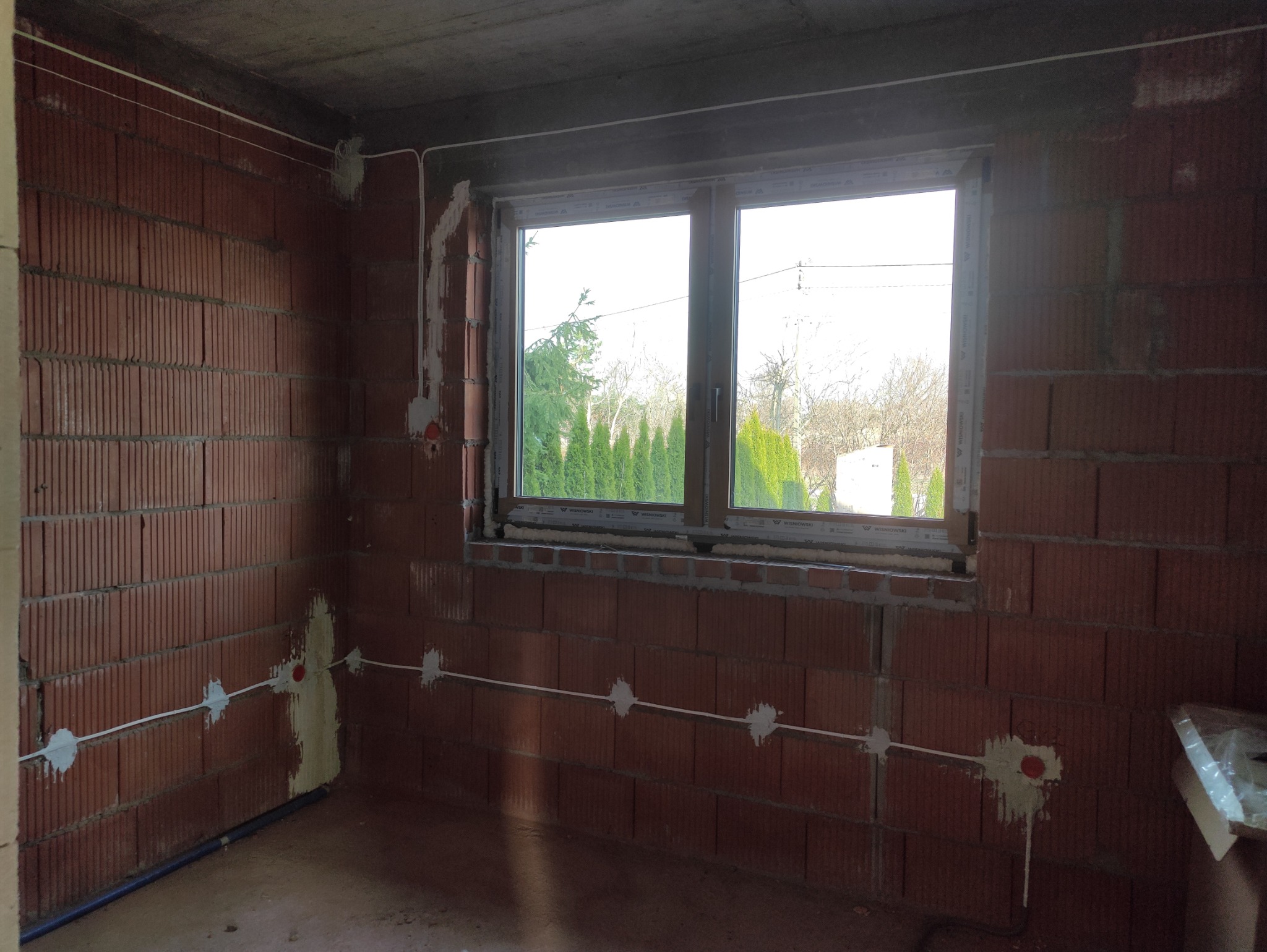 Wnętrze w trakcie prac elektrycznych: widoczne przewody rozprowadzone po ścianach z czerwonej cegły, puszki elektryczne, okno z widokiem na zieleń. Surowy stan wykończenia.