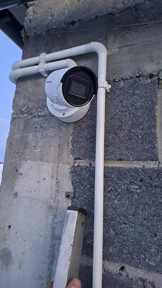 Zamontowana kamera monitoringu na szarej, betonowej ścianie. Białe rurki osłonowe prowadzą do kamery. Fragment drabiny w dolnej części kadru.