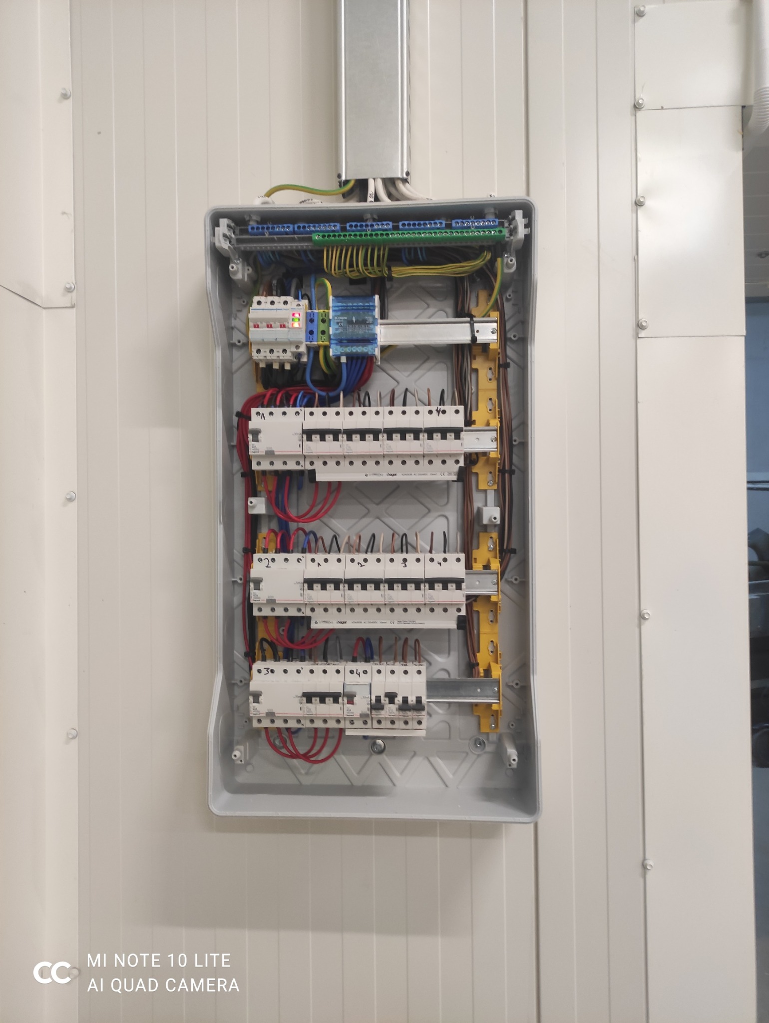 Otwarta skrzynka elektryczna z bezpiecznikami i okablowaniem na białej ścianie. Widoczne elementy instalacji elektrycznej wewnątrz obudowy.