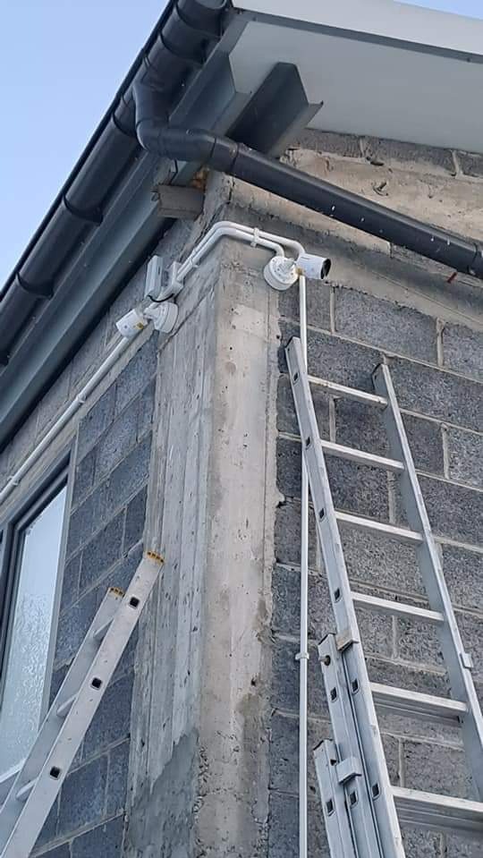 Dwie kamery monitoringu zamontowane na rogu budynku z szarej cegły, widoczne drabiny opierające się o ścianę. Instalacja zewnętrzna, białe przewody.