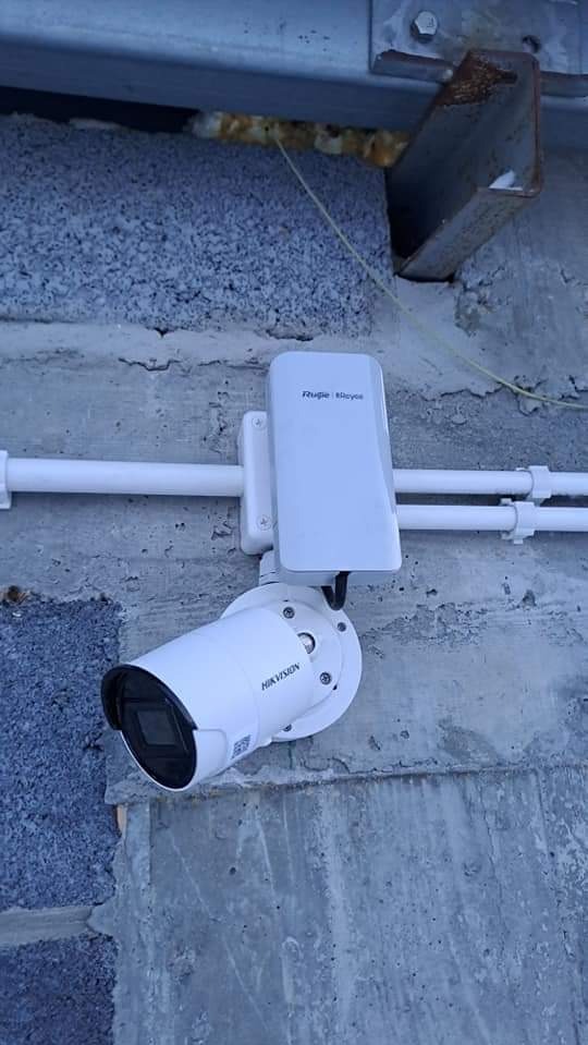 Biała kamera monitoringu Hikvision z anteną, zamontowana na szarej, betonowej ścianie. Widoczne przewody i elementy mocujące.